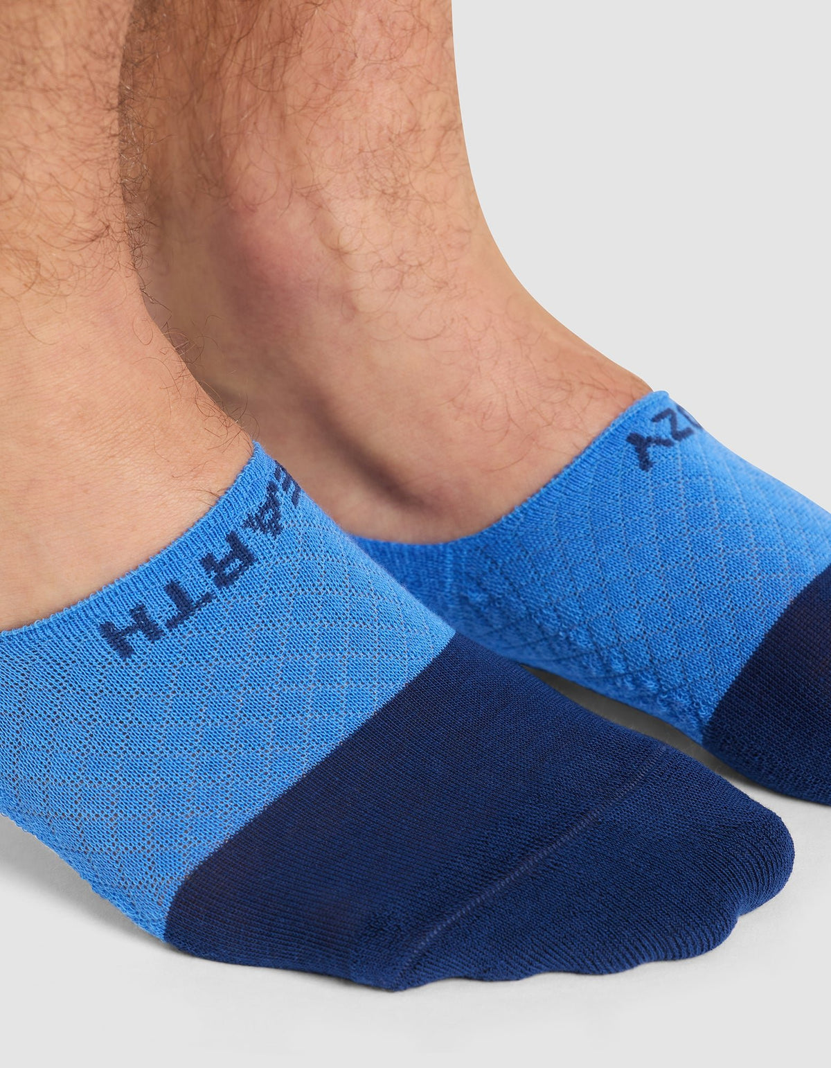 Close-up of two feet wearing Cozy Earth Essential No-Show Socks in blue, featuring a darker blue toe and "EARTH" near the ankle, shown on a light background.