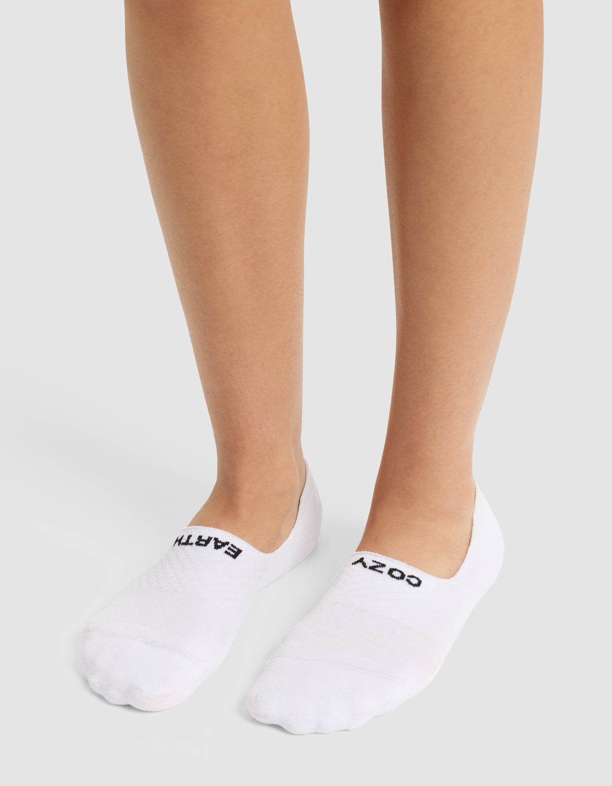 A person stands on a light surface wearing Cozy Earth’s Essential No-Show Sock, featuring "COZY" on the right sock and "EARTH" on the left.