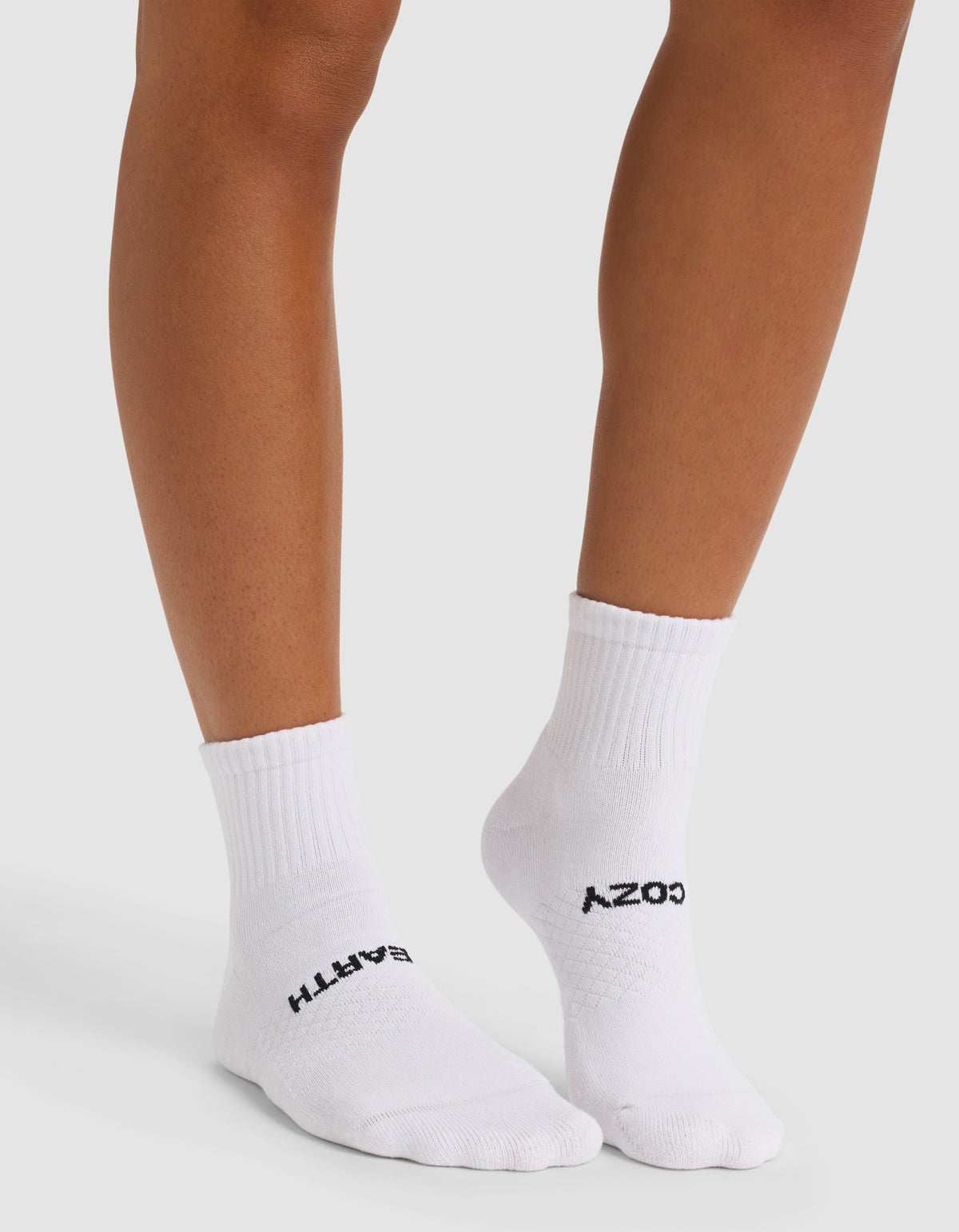 A person stands wearing Cozy Earth Essential Quarter Socks in white. "COZY" is written in black on the top of one sock and "EARTH" on the other. The background is plain and light-colored.