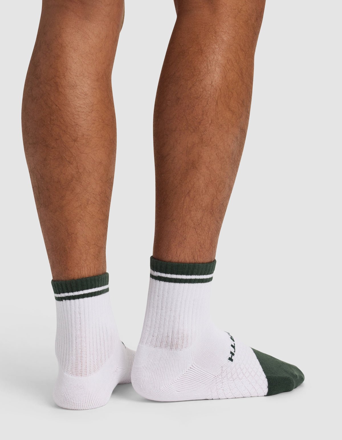 A person stands with their back to the camera, showing only their lower legs and feet in Cozy Earth’s Essential Quarter Sock - Last Chance: white socks with dark green cuffs and toes, ribbed texture, and a subtle sole pattern.