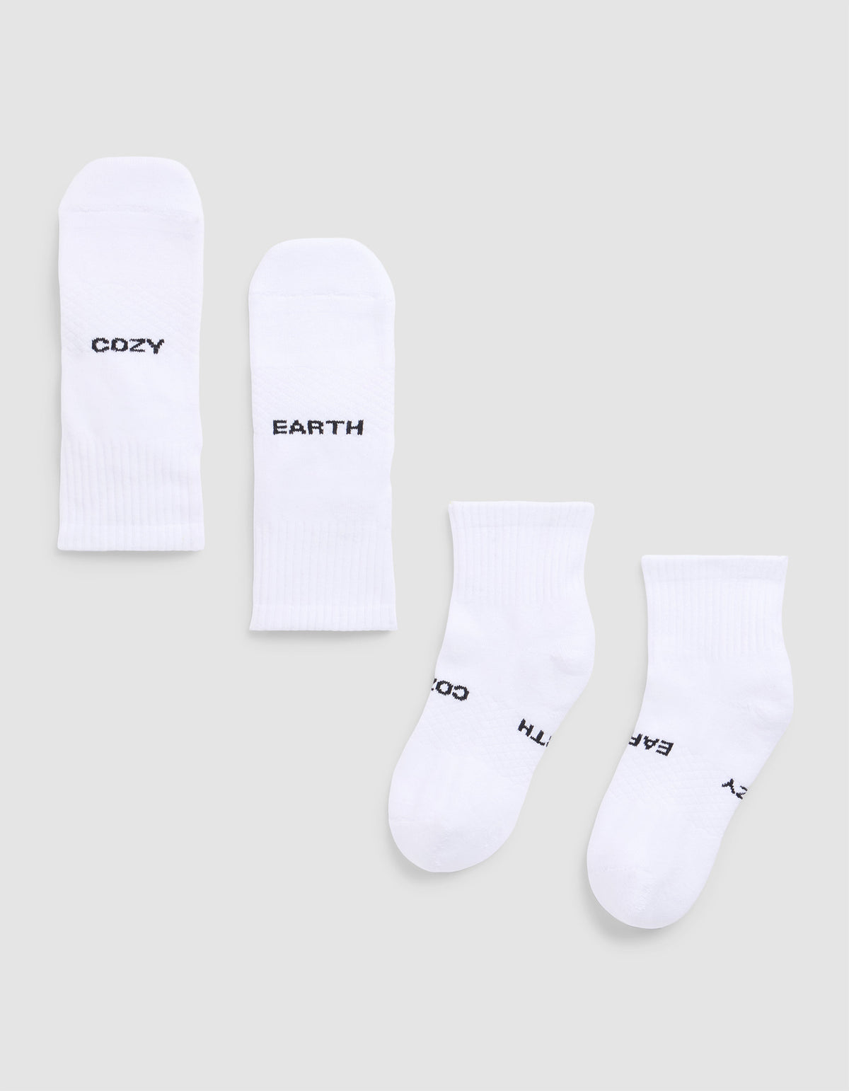 Four white Essential Quarter Socks from Cozy Earth are laid flat on a light gray background. Each features black text: one pair reads “COZY” and “EARTH,” while the other’s text shows parts of “EARTH,” “COZY,”.  