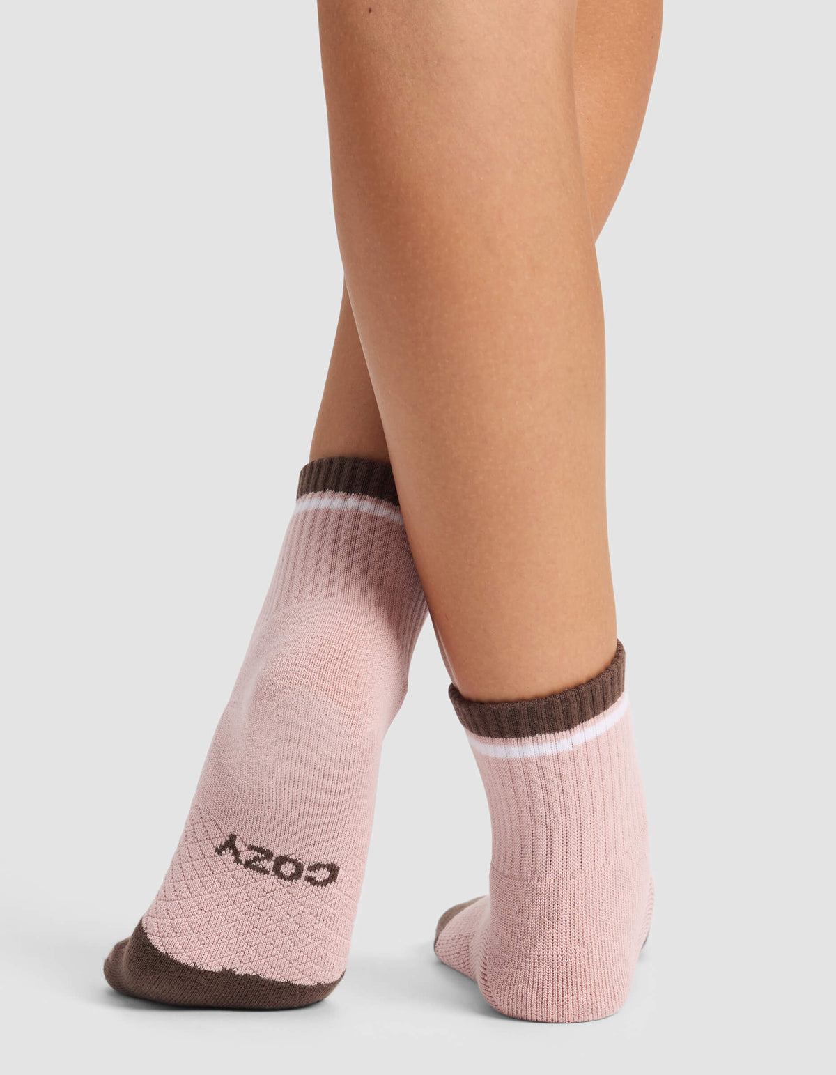 A person stands with feet crossed, wearing Cozy Earth Women's Essential Quarter Socks in light pink with brown heels, toes, and cuffs. "COZY" is printed in black on the bottom of one sock.