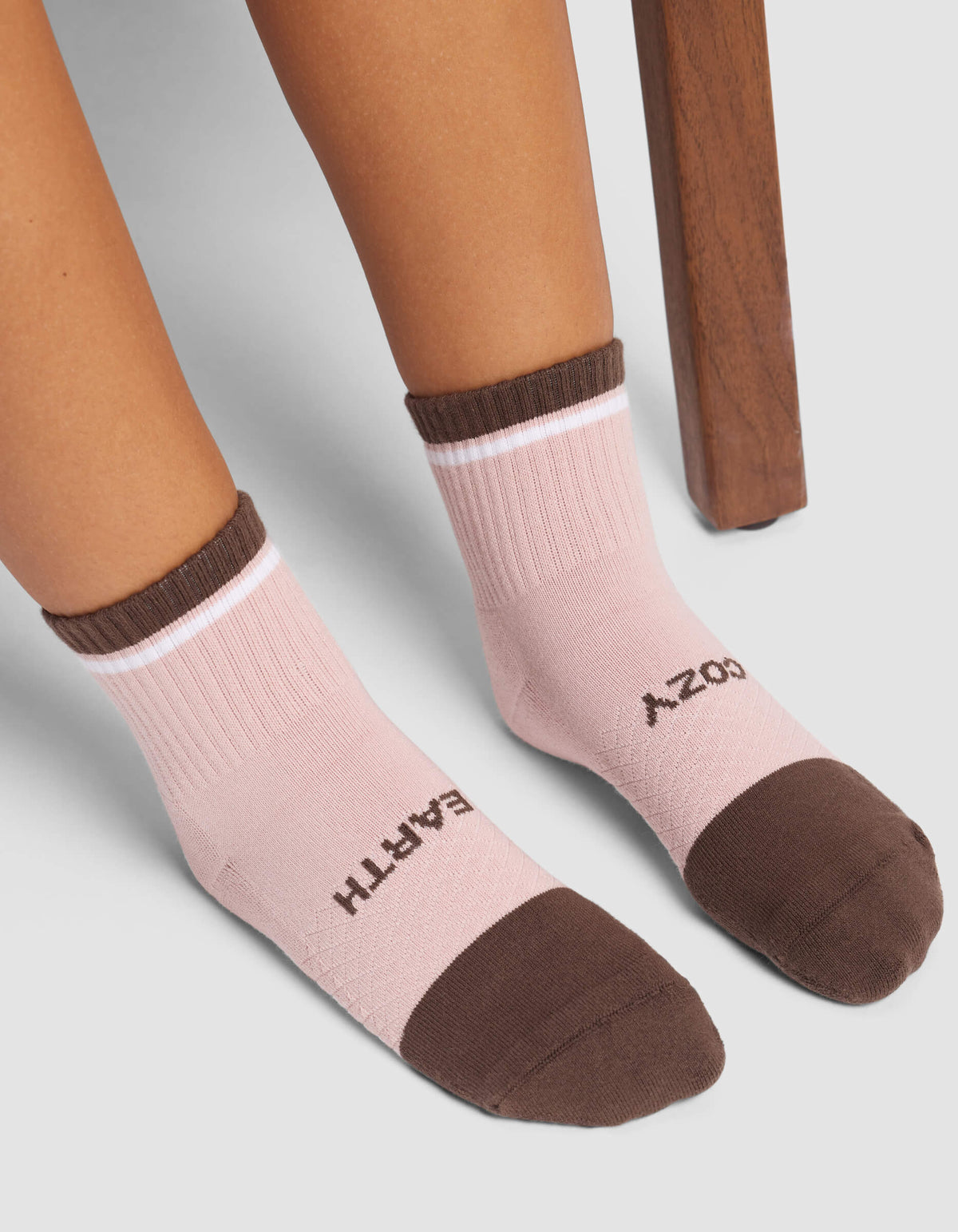 A person wearing Cozy Earth Women's Essential Quarter Socks in pink with brown toes and cuffs, featuring “COZY” on the right foot and “EARTH” on the left, sits near a wooden chair.