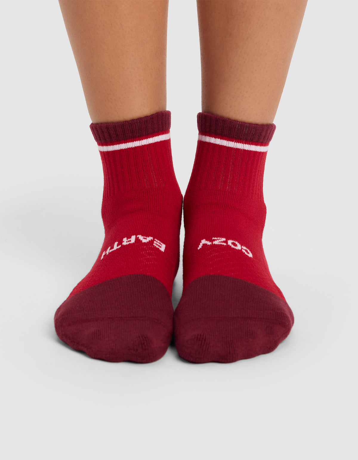 A pair of feet wearing Cozy Earth Essential Quarter Socks in red, featuring darker red toes and cuffs, white stripes near the top, and partially visible white text. The feet stand on a light grey surface.