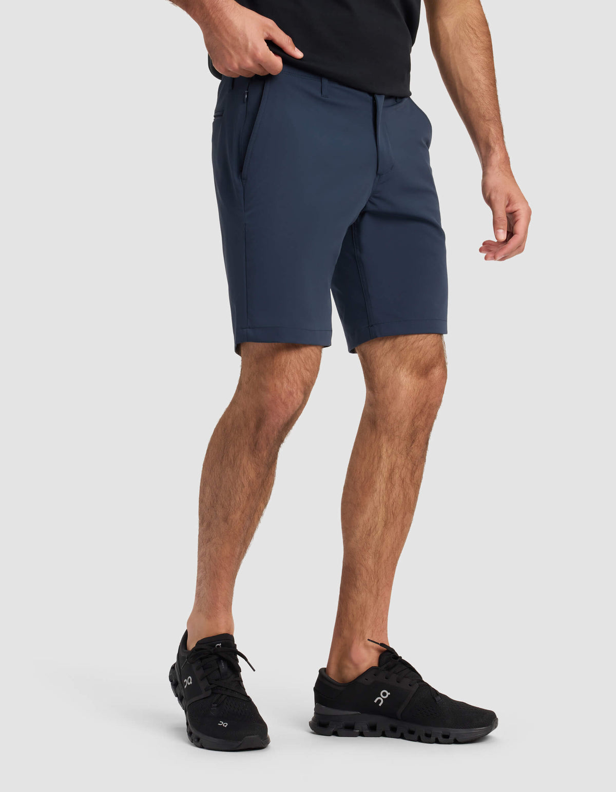 A person wearing a black t-shirt, Cozy Earth Men's Everywhere Short in navy, and black athletic shoes stands against a light gray background with one hand in their pocket.