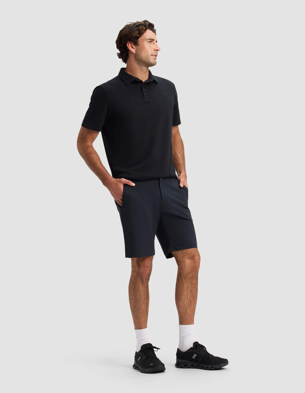 A man stands against a plain light gray background, wearing Cozy Earth's Men's Everywhere Short with his hands in his pockets, paired with a black polo shirt, white socks, and black sneakers as he looks to the side.