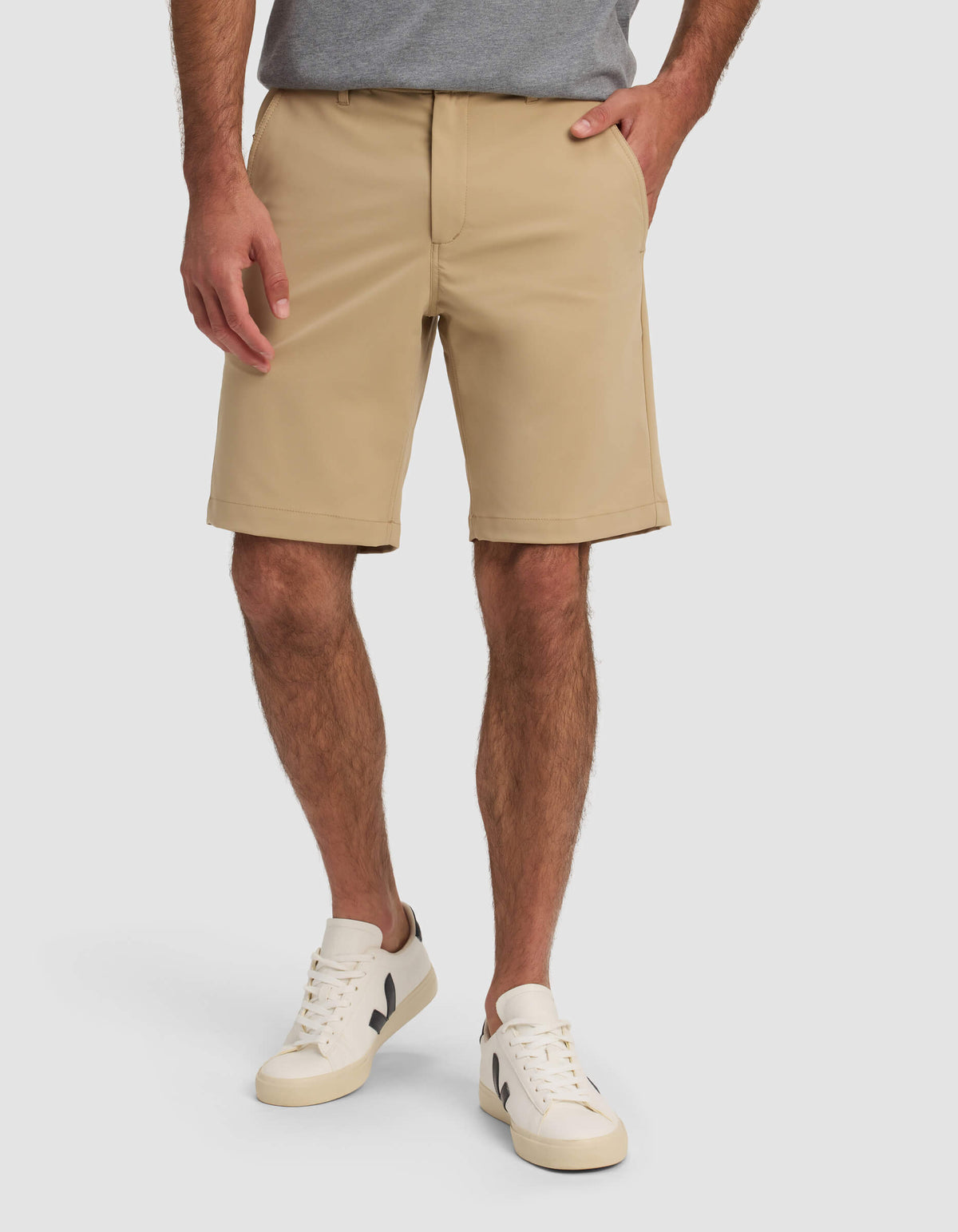 A man wearing Cozy Earth Men's Everywhere Short in beige, a gray t-shirt, and white sneakers with black accents stands with one hand in his pocket against a plain background.