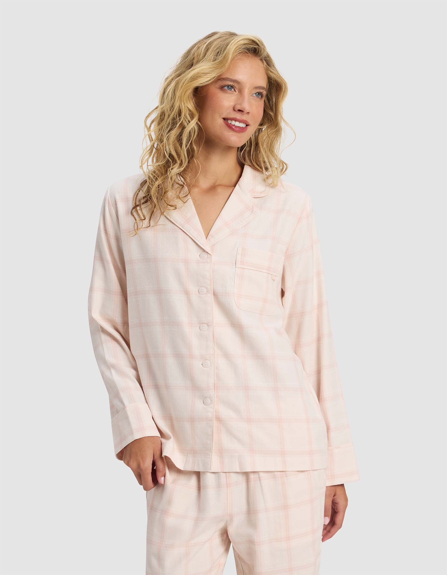 A woman with wavy blonde hair models the Cozy Earth Women's Farmhouse Long Sleeve Pajama Top, featuring a light pink plaid design, button-up front, and matching pants, standing against a plain light background and smiling slightly.|Color:Quartz Plaid