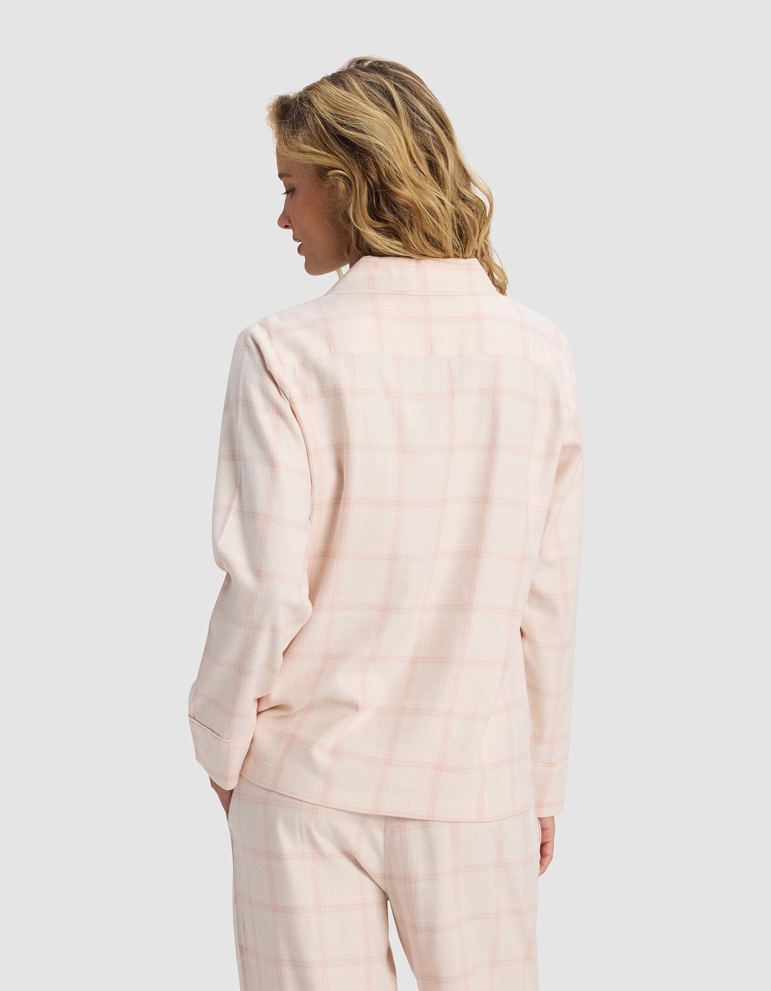 A woman with wavy blonde hair is seen from the back, wearing the Cozy Earth Women's Farmhouse Long Sleeve Pajama Top in pale pink plaid, paired with matching pants, against a plain light gray background.|Color:Quartz Plaid
