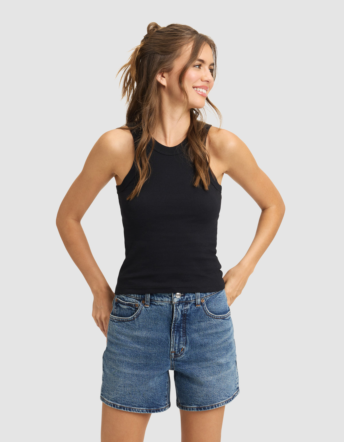 A woman with long brown hair smiles and looks aside, wearing the Cozy Earth Women's Fine Ribbed Tank in black and blue denim shorts. She stands with hands in pockets against a plain light background.