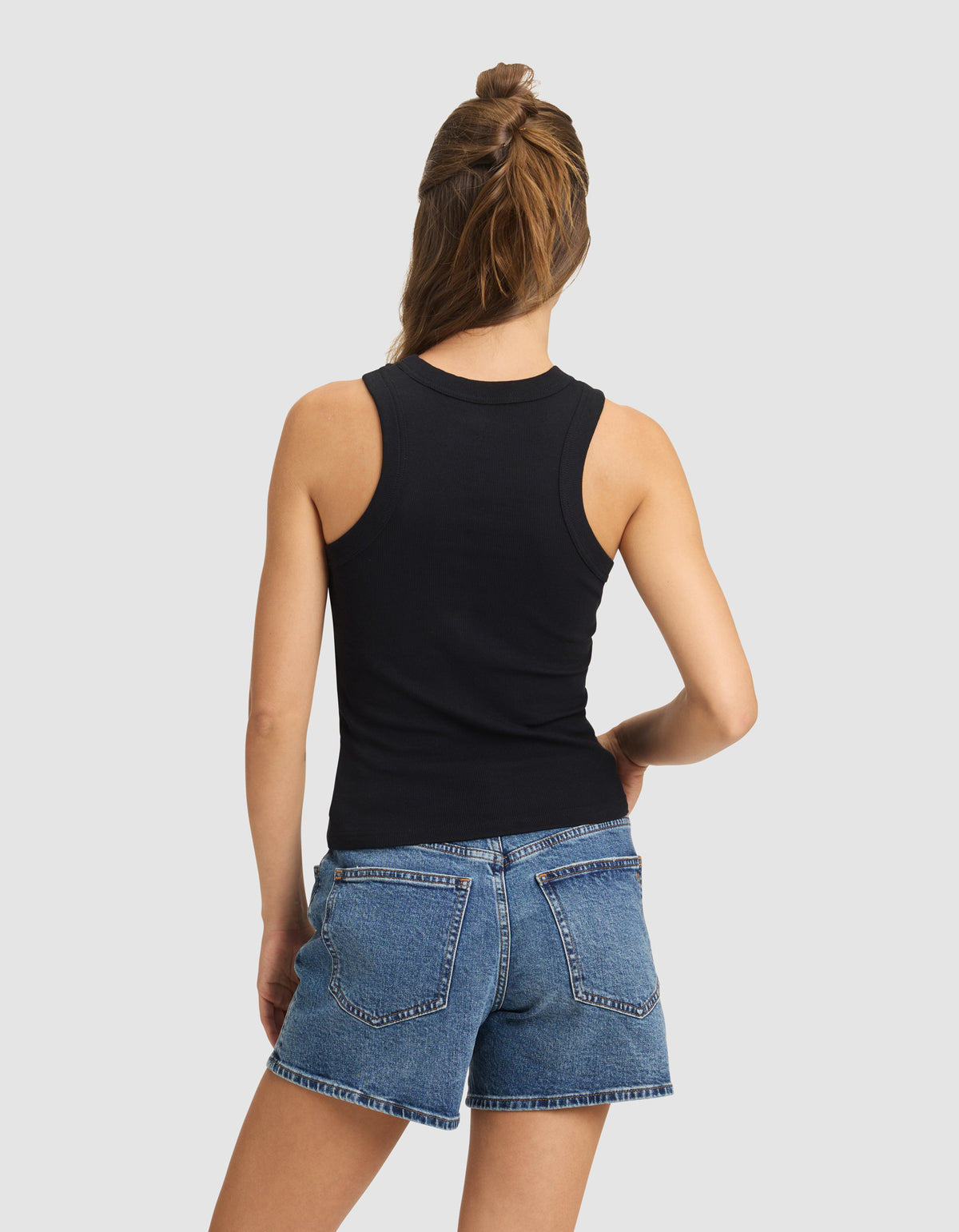 A woman with light brown hair wears the Cozy Earth Women's Fine Ribbed Tank in black, paired with blue denim shorts. She stands facing away from the camera, hand on hip, against a plain light gray background.
