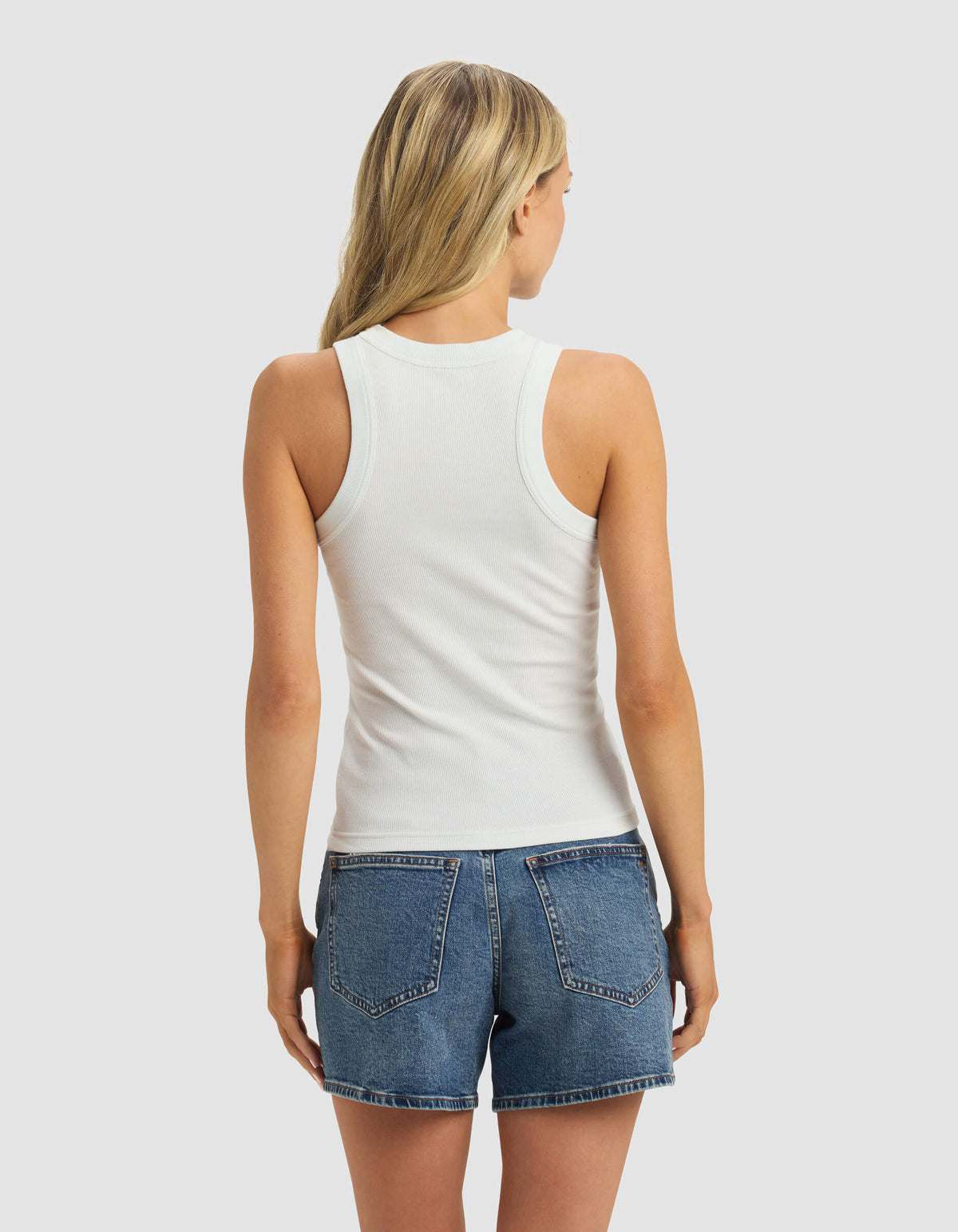 A woman with long blonde hair is seen from the back wearing the Cozy Earth Women's Fine Ribbed Tank in white and blue denim shorts, set against a light gray background.