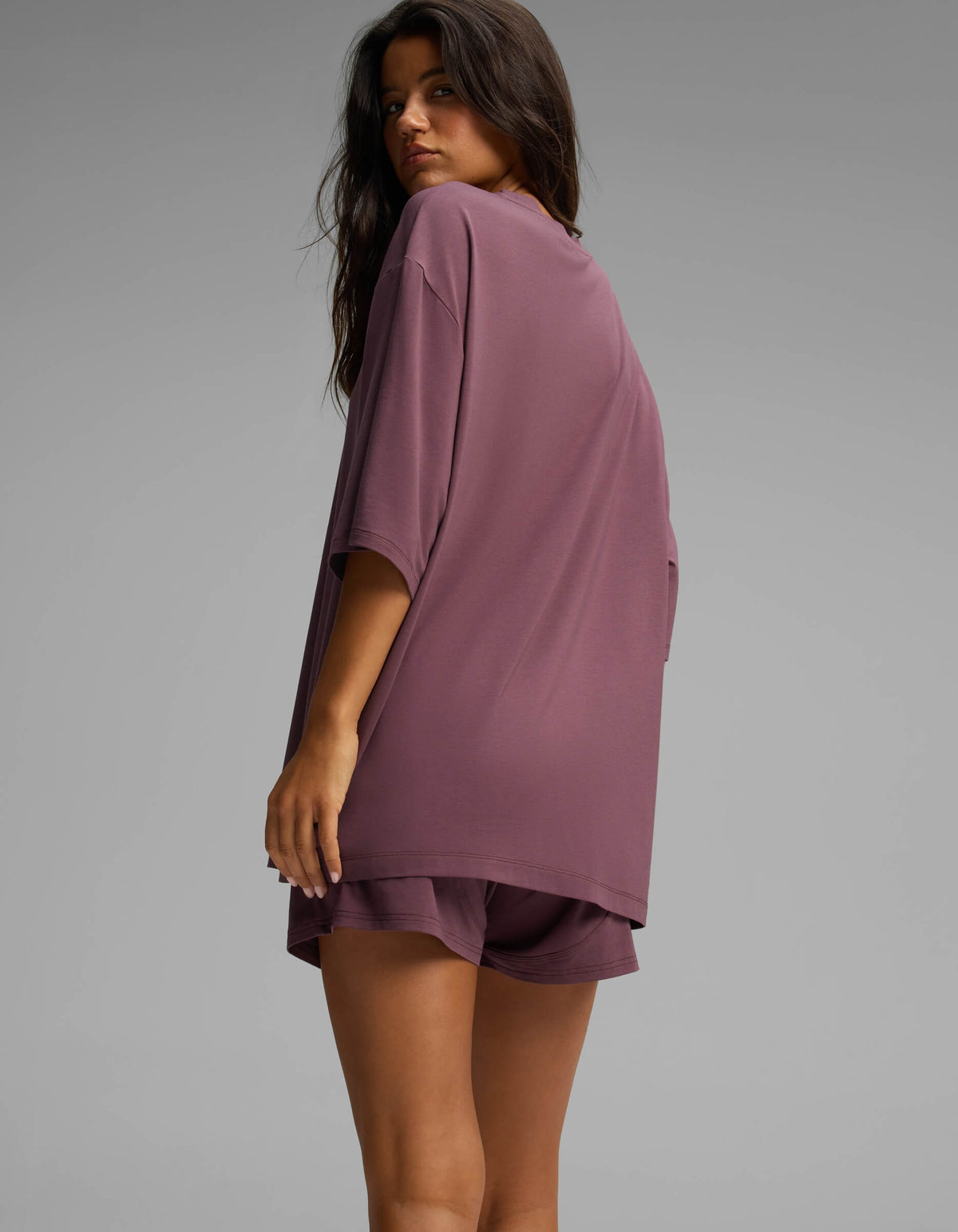 A woman with long hair stands sideways and looks over her shoulder against a plain gray background, wearing the Cozy Earth Women’s First Layer Boyfriend Sleep Tee in mauve.