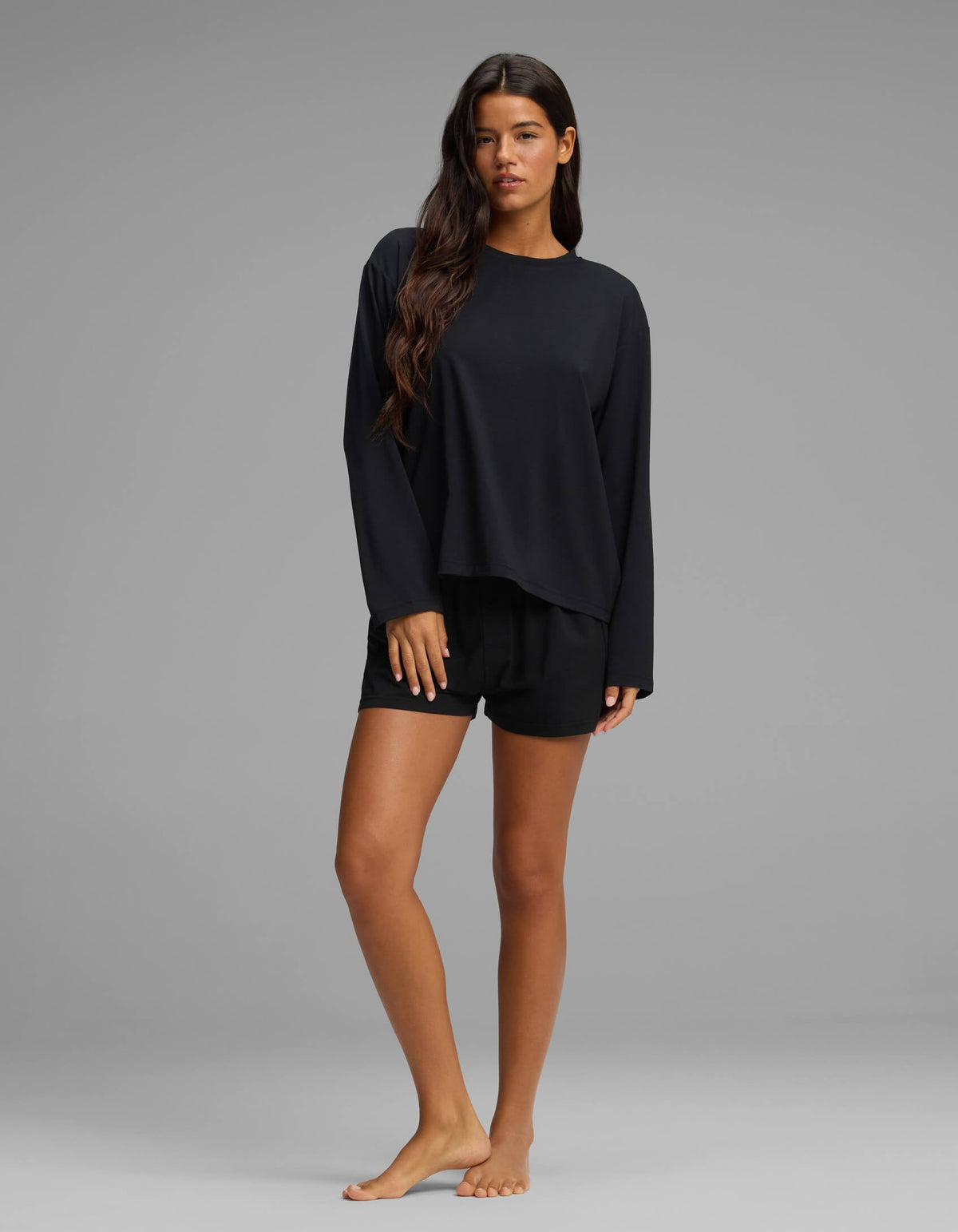 A woman with long brown hair stands barefoot on a grey background, modeling the Cozy Earth Women’s First Layer Long Sleeve Sleep Tee & Boxer Short Set in black, looking at the camera with a neutral expression.