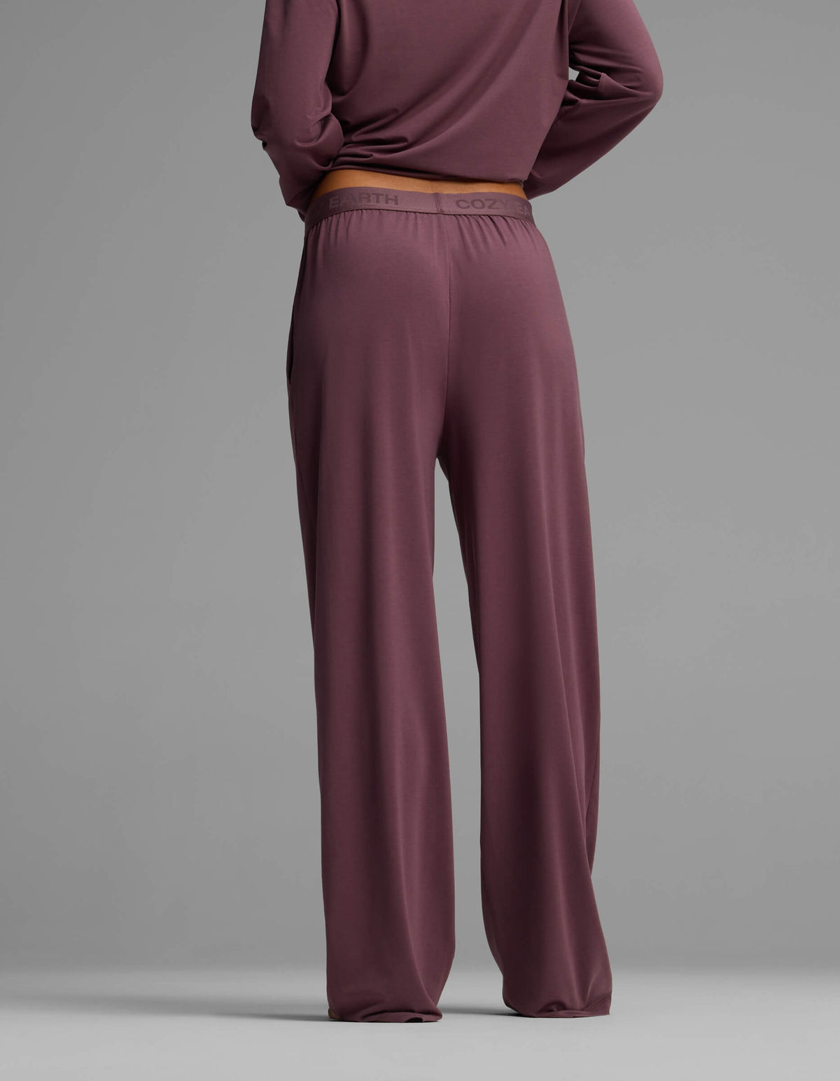 A woman is seen from the back wearing Cozy Earth’s Women’s First Layer Pajama Pant in a loose mauve fit, paired with a matching long-sleeve top, set against a plain gray background.