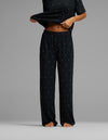 A person stands barefoot against a plain gray background, wearing the Cozy Earth Women’s First Layer Pajama Pant and a matching loose black top with small white polka dots, lifting the shirt slightly to reveal their midriff.|Color:Mini Ghost