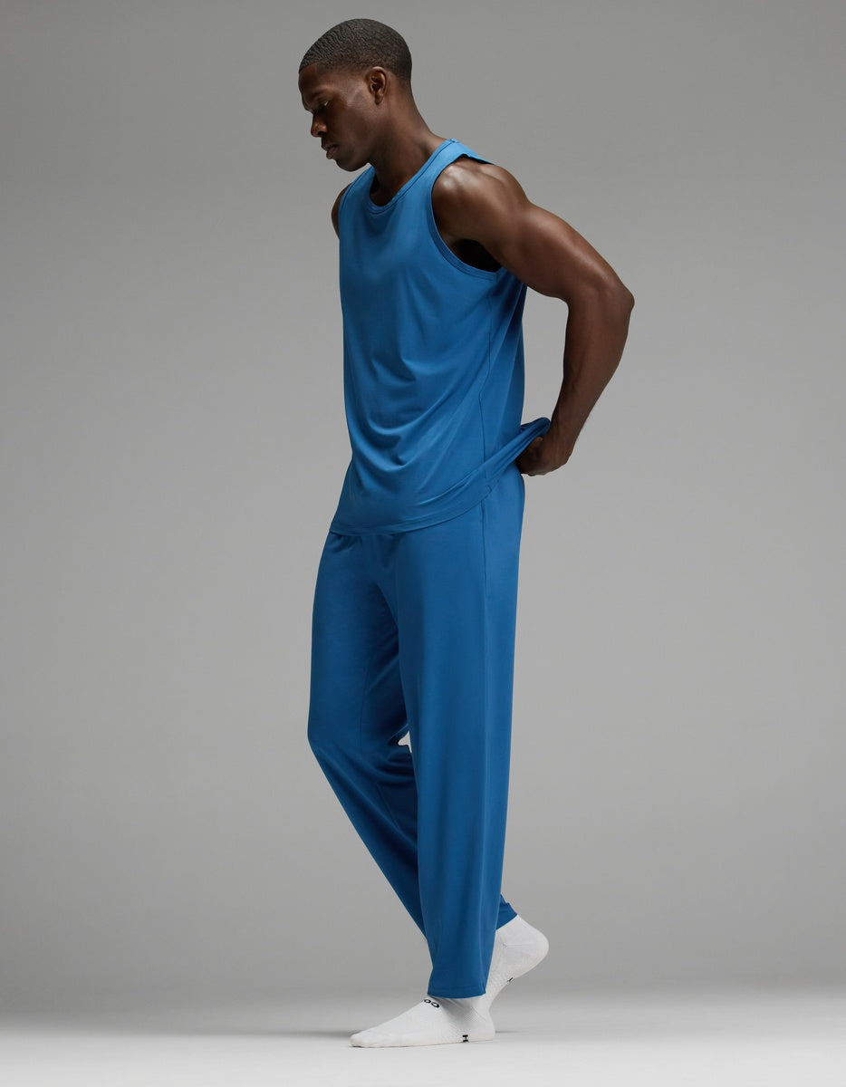 Wearing Cozy Earth’s Men’s First Layer Pajama Tank & Pant Set in blue, a person stands against a plain gray background with their hands behind their back, looking down. They complete the look with white socks.|Color:Anchor