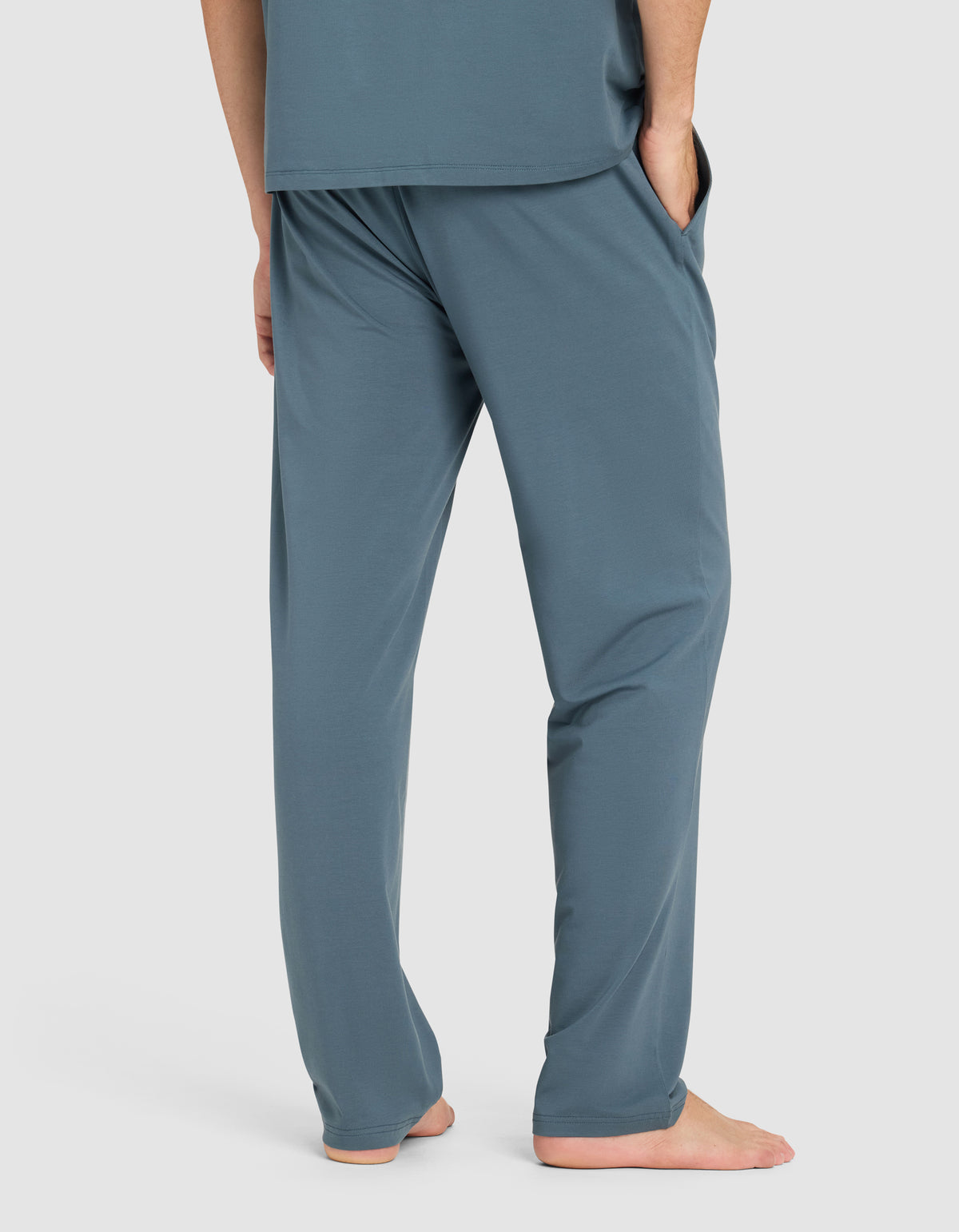 A man stands barefoot with hands in pockets, wearing Cozy Earth's Men's First Layer Pajama Pant in teal blue, paired with a matching top. He is shown from the waist down against a plain light background.