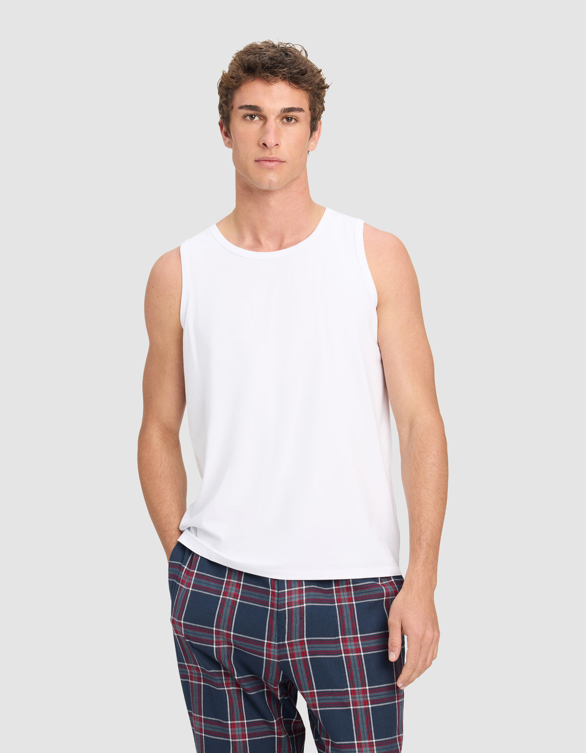 A man wears the Cozy Earth Men's First Layer Pajama Tank in white, paired with blue plaid pants, standing against a light gray background and looking directly at the camera with a neutral expression.|Color:White