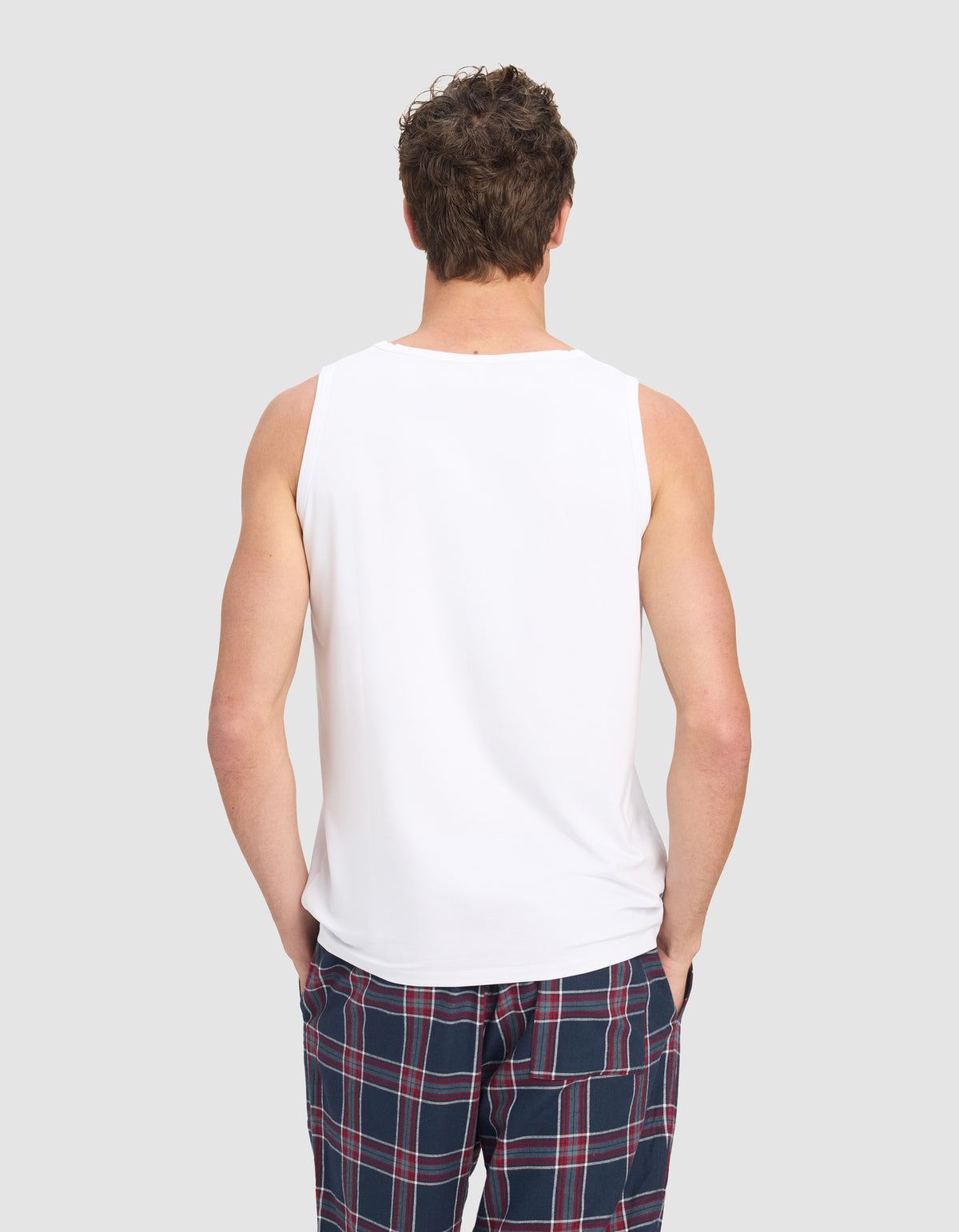 A man models the Cozy Earth Men's First Layer Pajama Tank in white with blue plaid pajama pants featuring red and white stripes, standing against a plain light gray background.