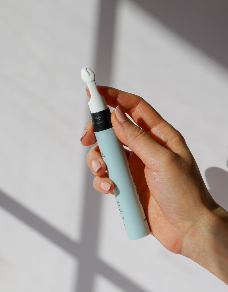 A hand with neatly manicured nails holds a light blue tube of Cozy Earth Eye Cream with a white, rounded applicator tip, set against a softly lit background featuring shadow patterns.