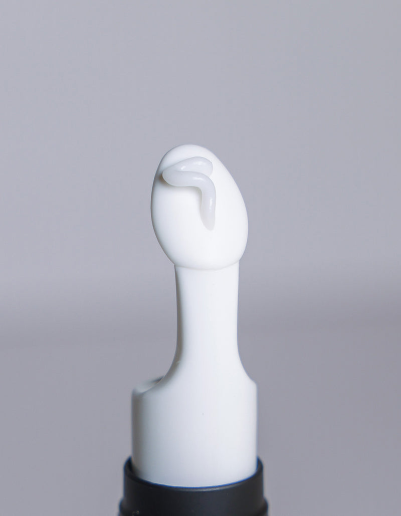 A close-up of a Cozy Earth Eye Cream white plastic applicator tip with a small amount of the cream on it, set against a plain, light gray background.