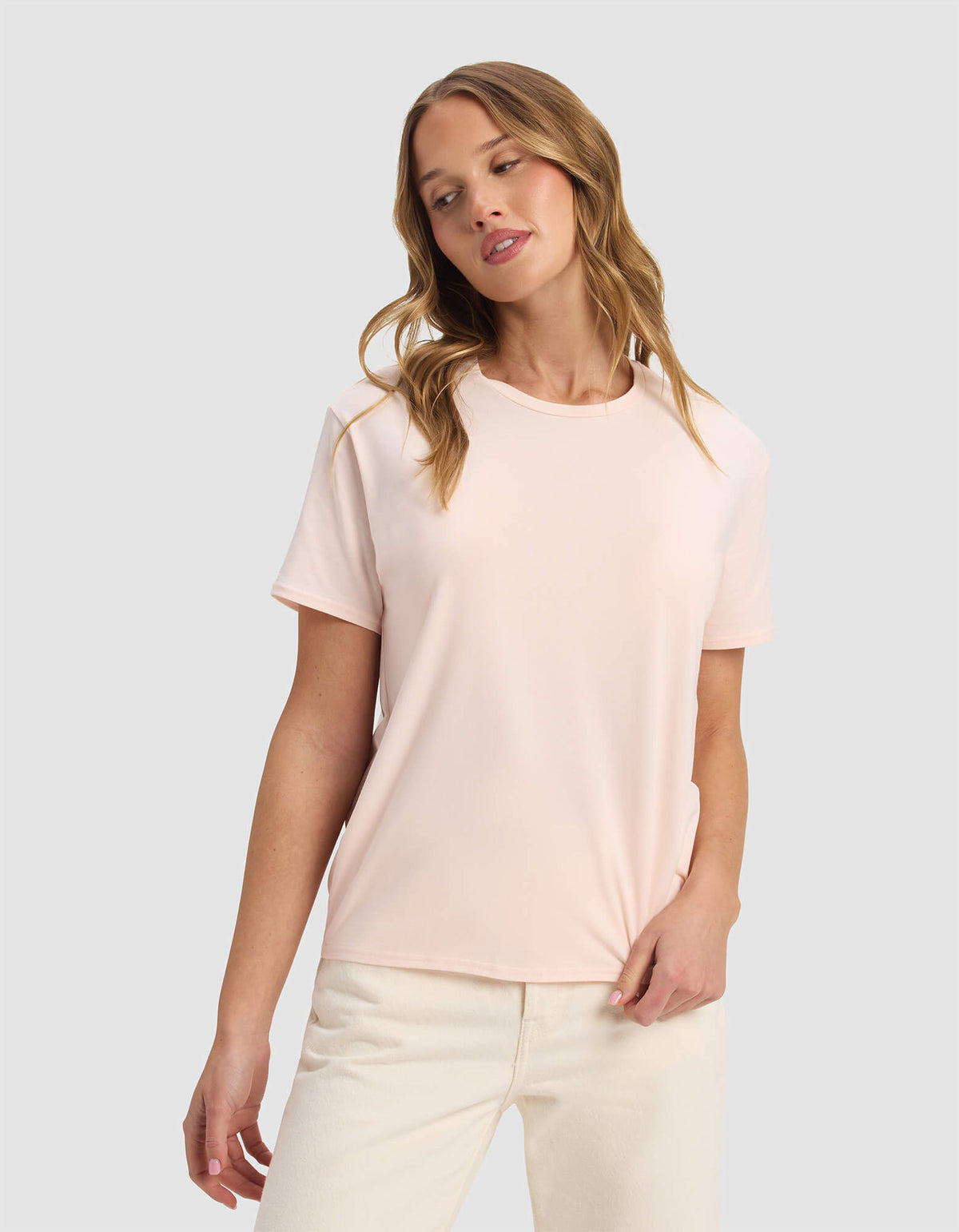 A woman with wavy blonde hair wears the Cozy Earth Women's Go-To Tee in Blossom and off-white pants, standing against a plain light gray background with a relaxed expression, looking slightly to the side.|Color:Blossom