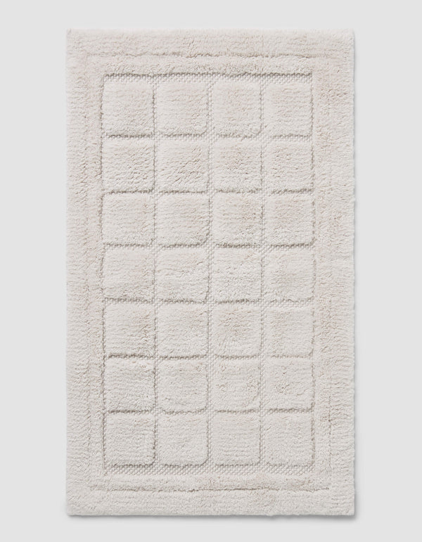The Cozy Earth Grid Bath Mat is a rectangular, off-white mat with a raised grid design and a thick, fluffy border.|Color:Light Grey