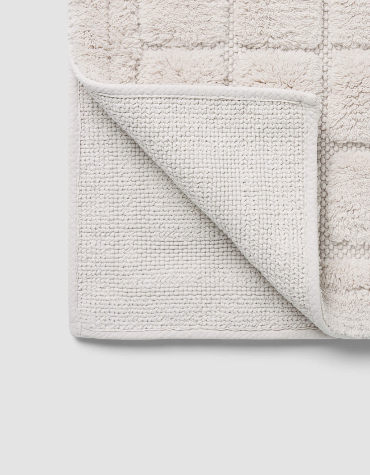 A close-up of the Cozy Earth Grid Bath Mat shows its light color, textured grid pattern, and one corner flipped to reveal the woven underside.