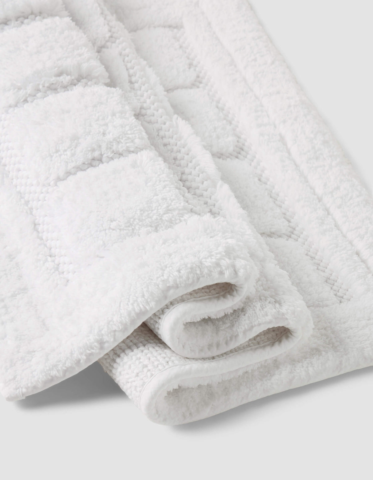 A close-up of the Folded Grid Bath Mat by Cozy Earth, featuring a white textured geometric pattern, displayed on a light gray background.|Color:White