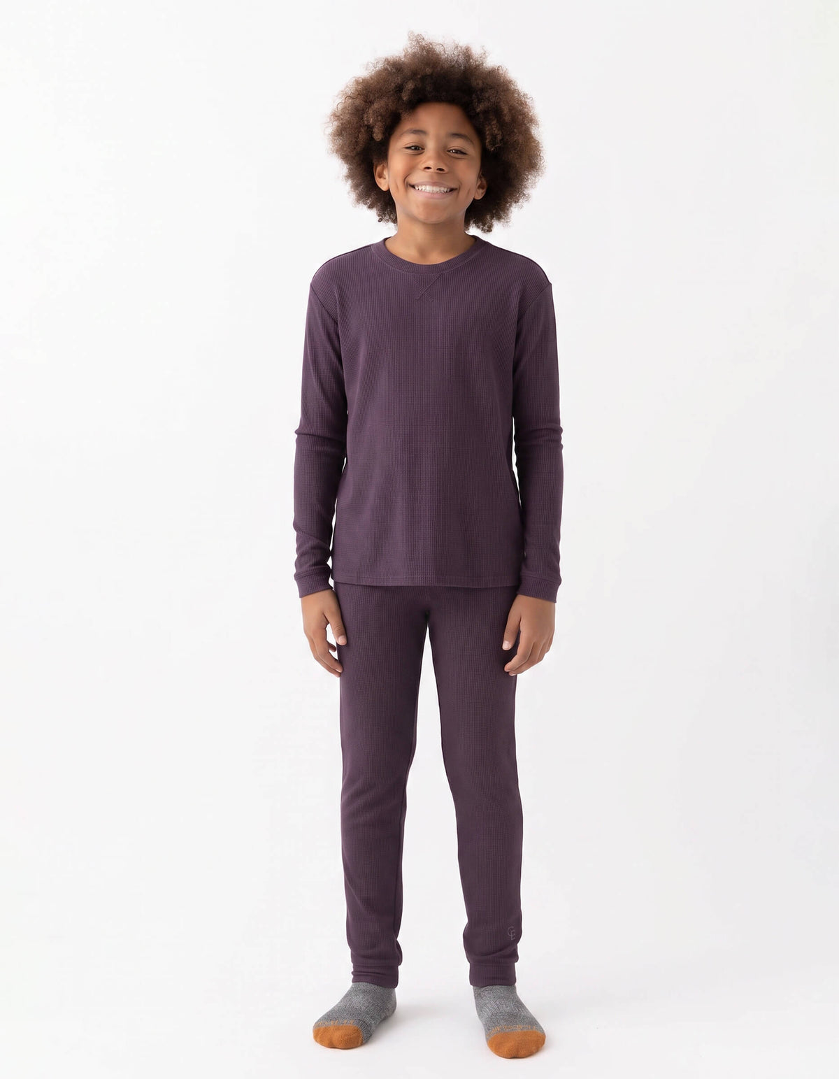 A smiling child with curly hair stands against a white background, wearing the Cozy Earth Children's Bamboo Waffle Knit Base Layer Set - Last Chance, paired with gray and orange socks.