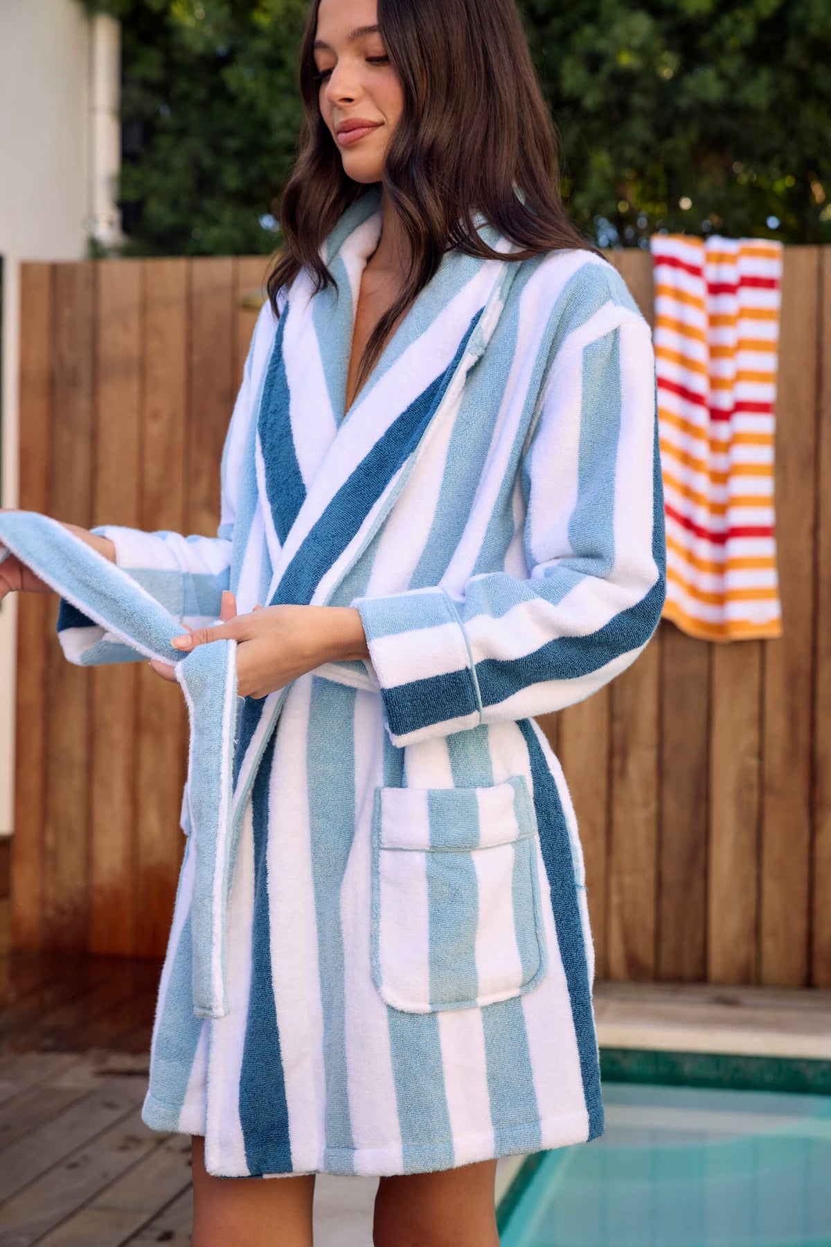 A woman in the Cozy Earth Classic Stripe Resort Robe stands by a pool, adjusting the belt. A yellow and white striped towel hangs on a wooden fence behind her, with greenery in the background.