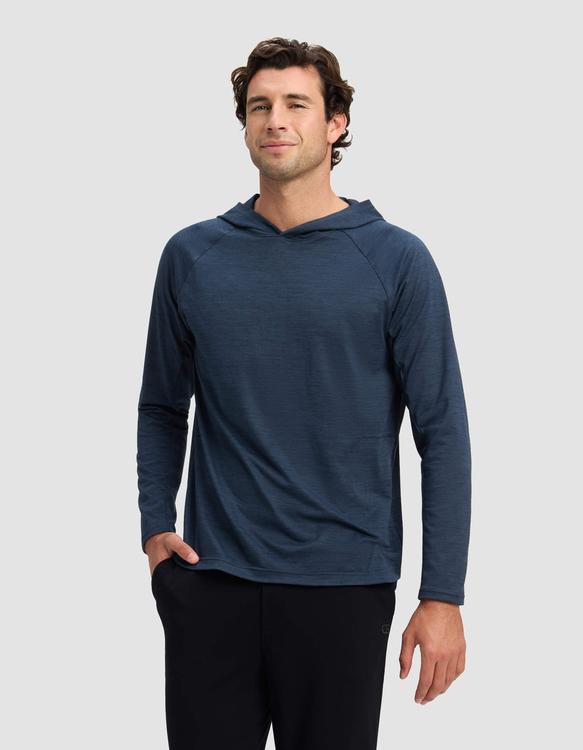 A man wearing Cozy Earth's Men's Lightweight Performance Hoodie in dark blue and black pants stands against a plain light gray background, one hand in his pocket and a slight smile.