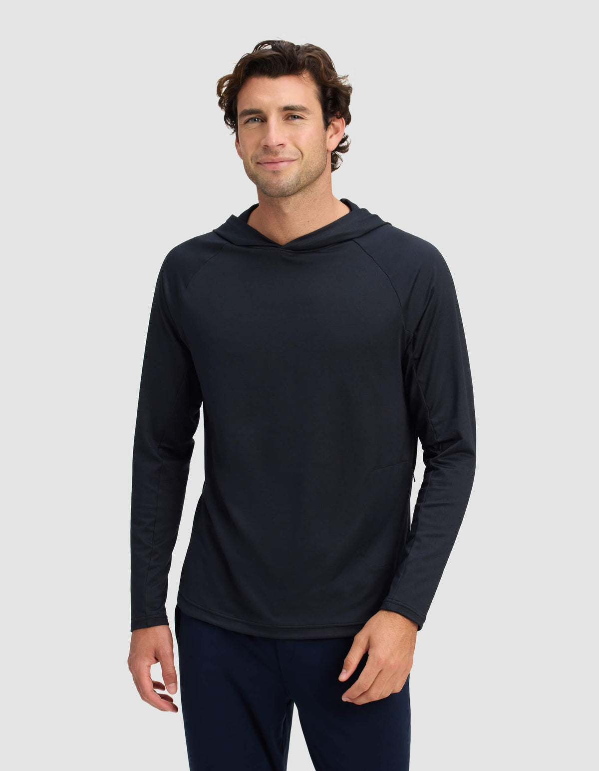 A man with short, wavy brown hair is wearing the Men's Lightweight Performance Hoodie by Cozy Earth, standing against a plain light gray background and smiling slightly at the camera.