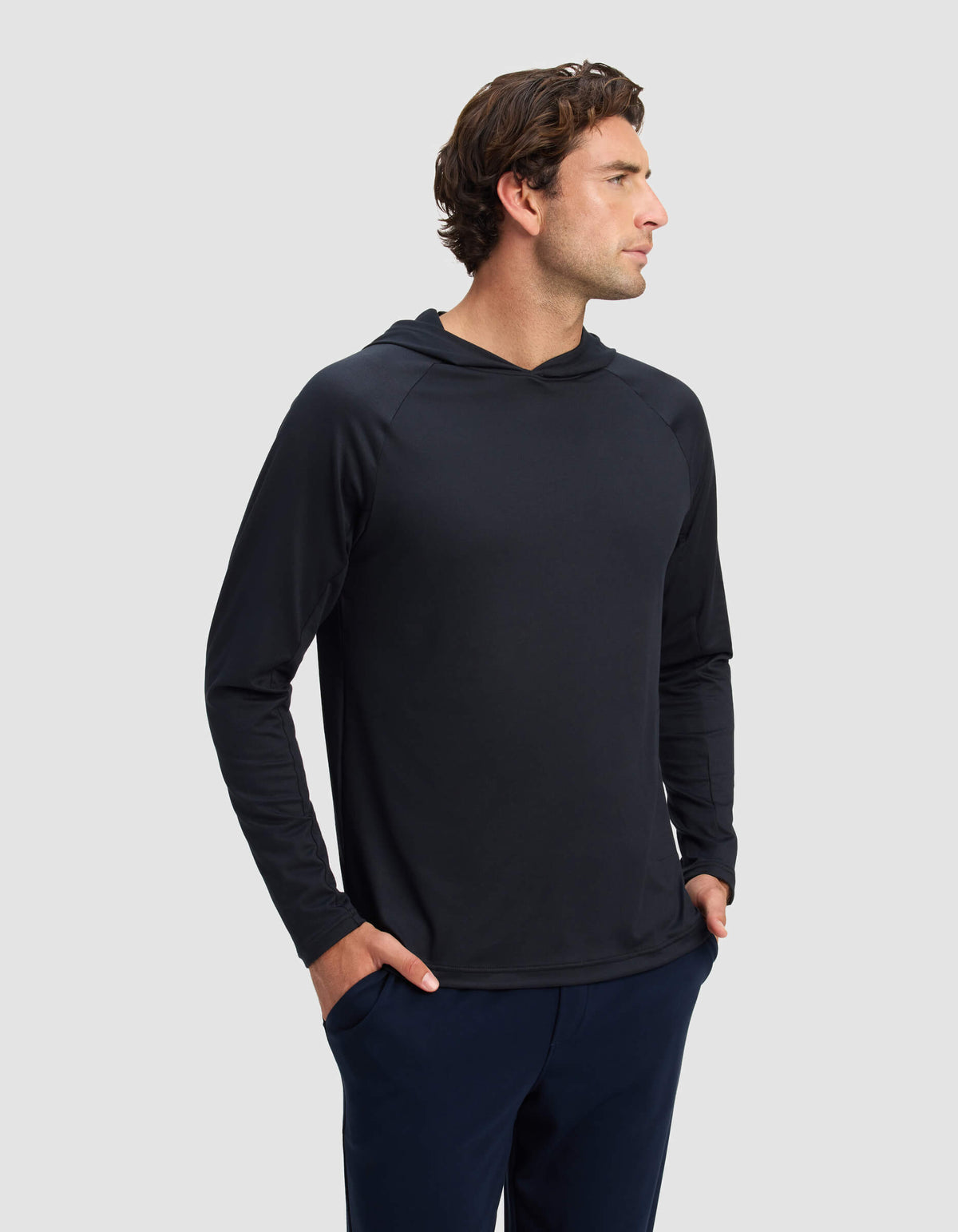 A man with wavy brown hair, wearing the Cozy Earth Men's Lightweight Performance Hoodie and dark pants, stands with his hands in his pockets, looking to the side against a plain light gray background.|Color:Jet Black