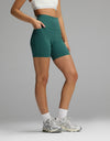 A person wearing Women’s Liv Biker Short 6″ with Pockets in teal with side pockets, a white sports bra, white socks, and silver athletic sneakers.|Color:Uniform