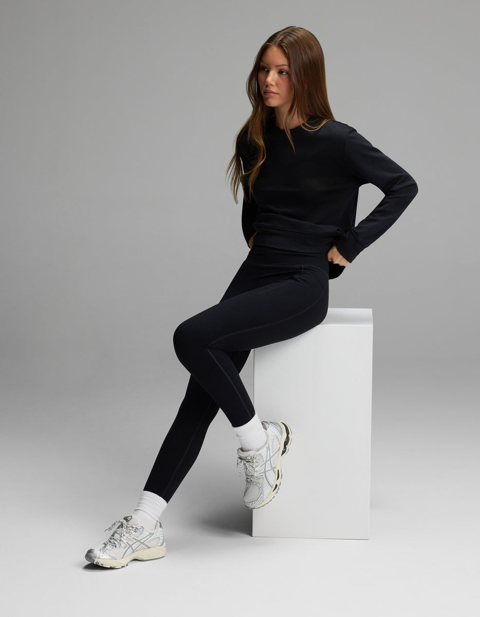A person wearing a black long-sleeve shirt and Women’s Liv Leggings with Pockets, paired with white socks and white sneakers, sitting on a white pedestal.