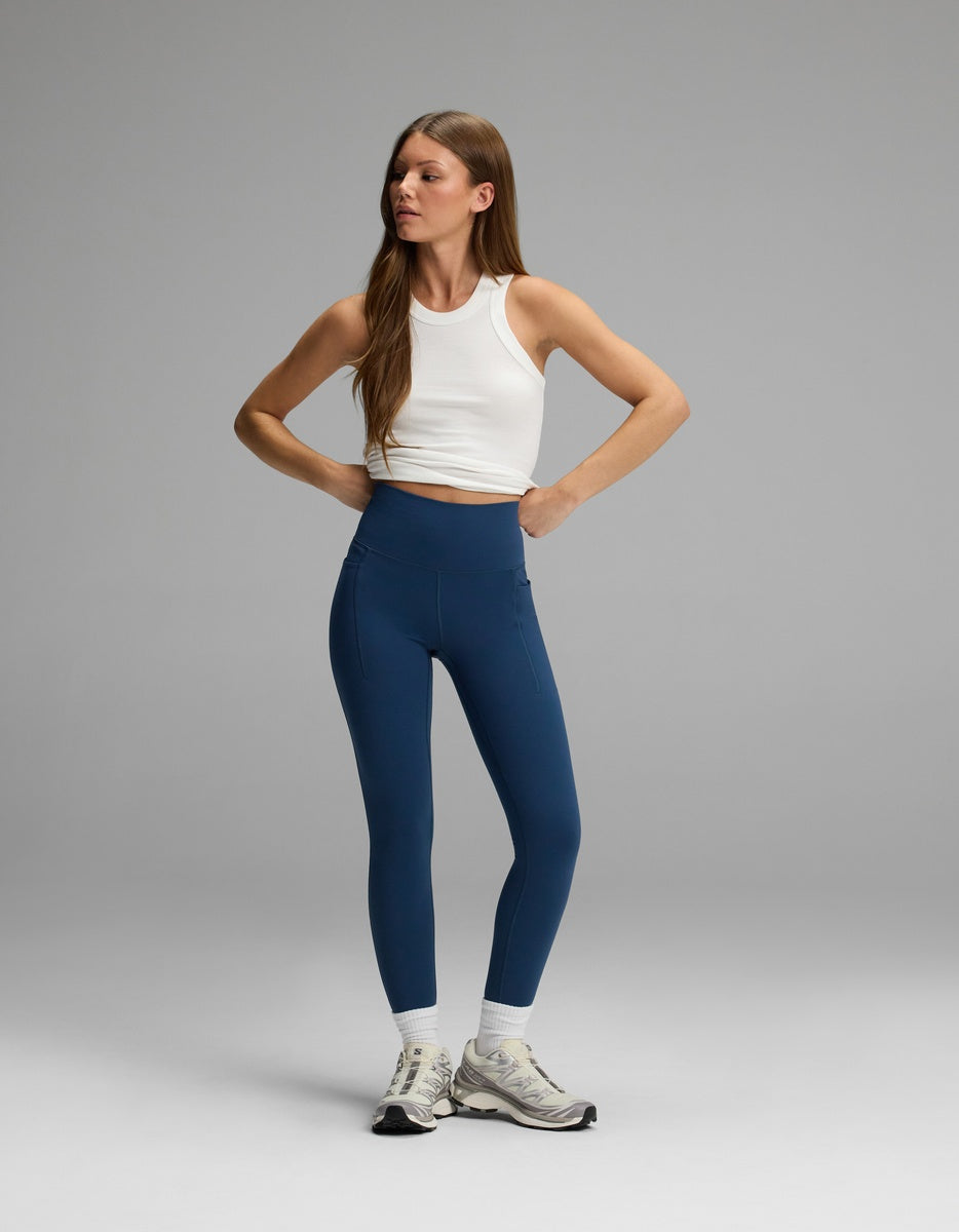 A woman wearing a white tank top and blue Women’s Liv Leggings with Pockets that feature a high-waisted fit.|Color:Obsidian