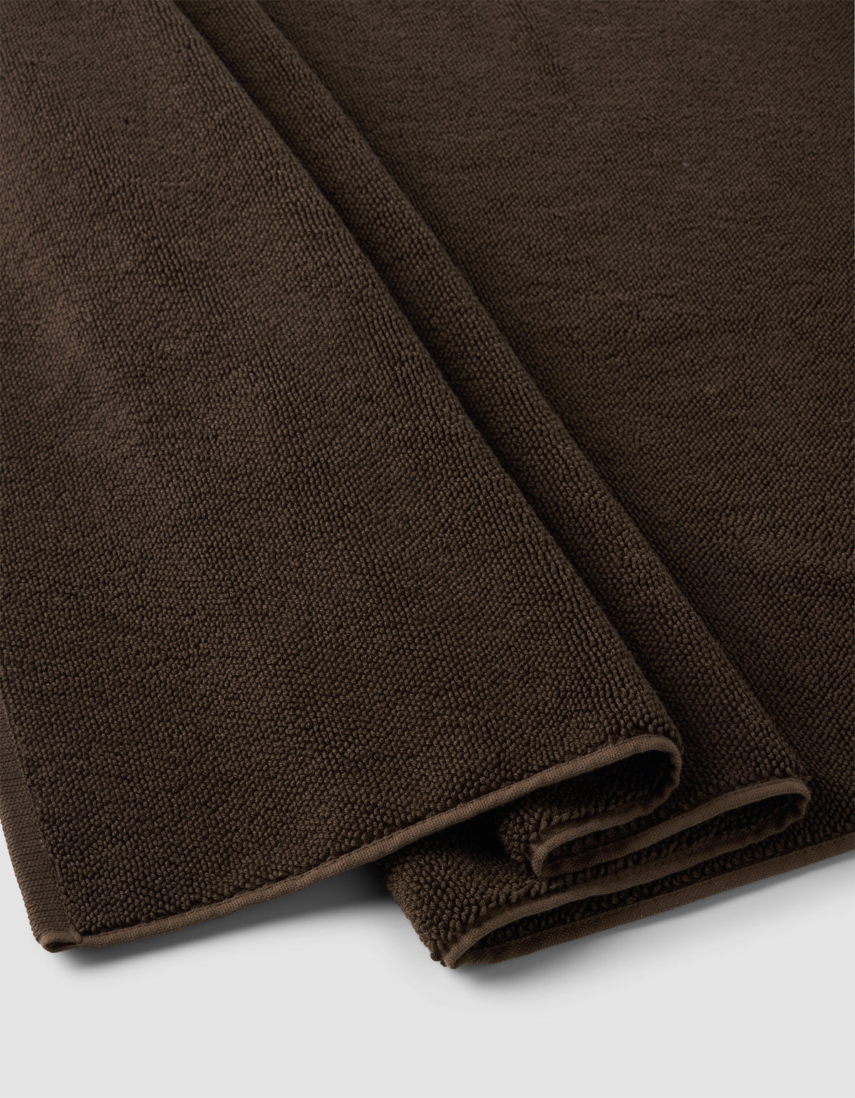 A close-up of two folded Cozy Earth Loop Terry Bath Mats in dark brown, featuring textured woven fabric, placed on a light gray surface.