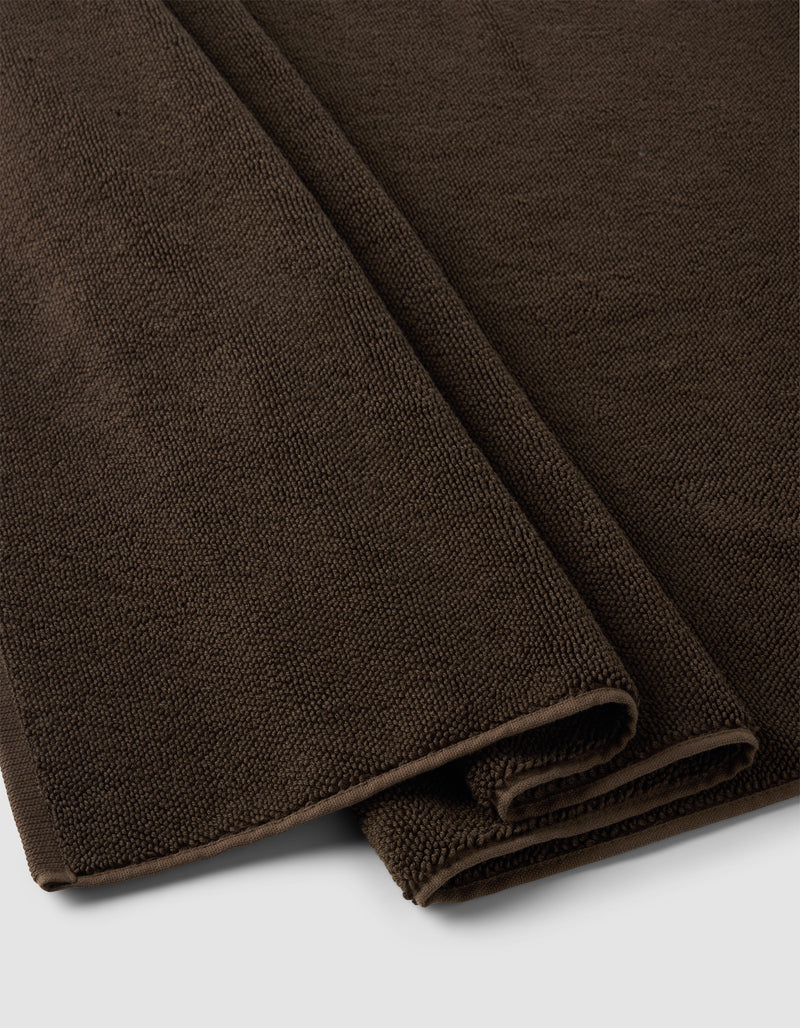 A close-up of two folded Cozy Earth Loop Terry Bath Mats in dark brown, featuring textured woven fabric, placed on a light gray surface.