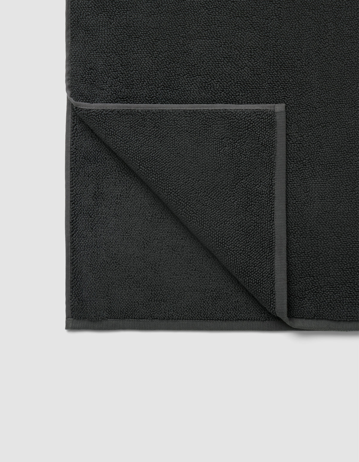 The Cozy Earth Loop Terry Bath Mat in dark gray is pictured neatly folded at one corner, displaying its textured surface and smooth edge border on both front and back sides.