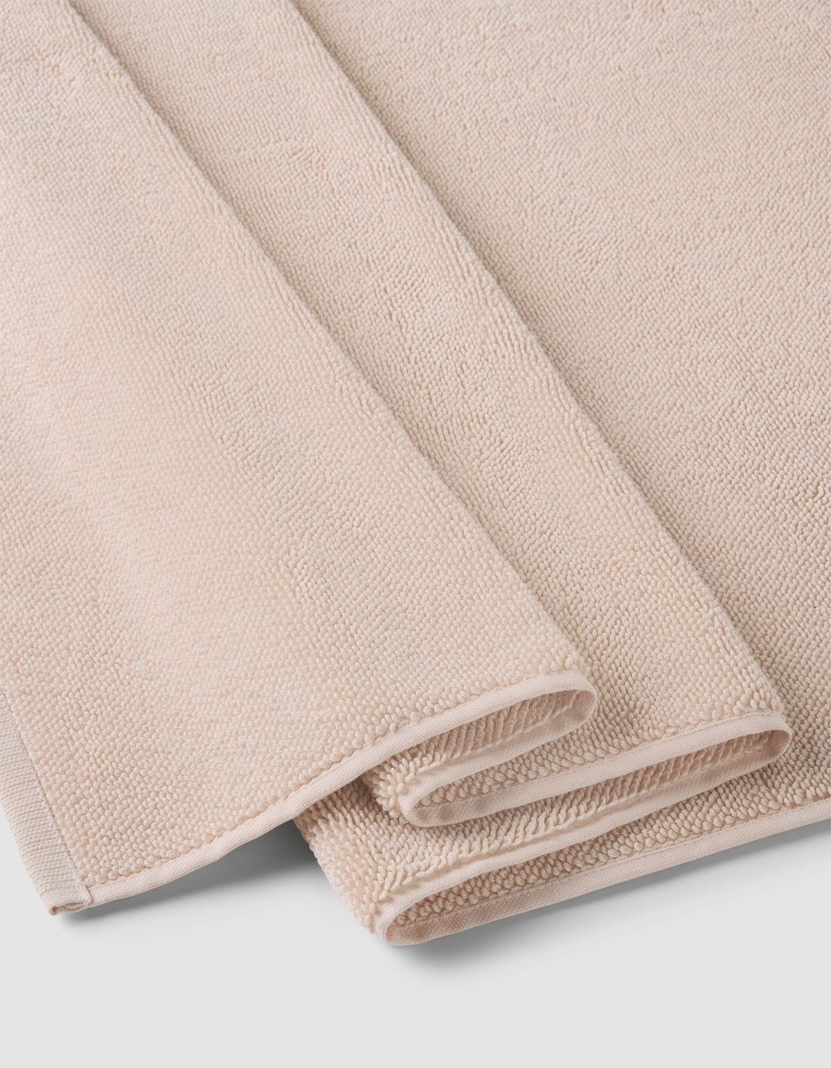 A Loop Terry Bath Mat by Cozy Earth, in light beige with a soft, textured surface, is neatly folded and stacked on a white background.|Color:Petal