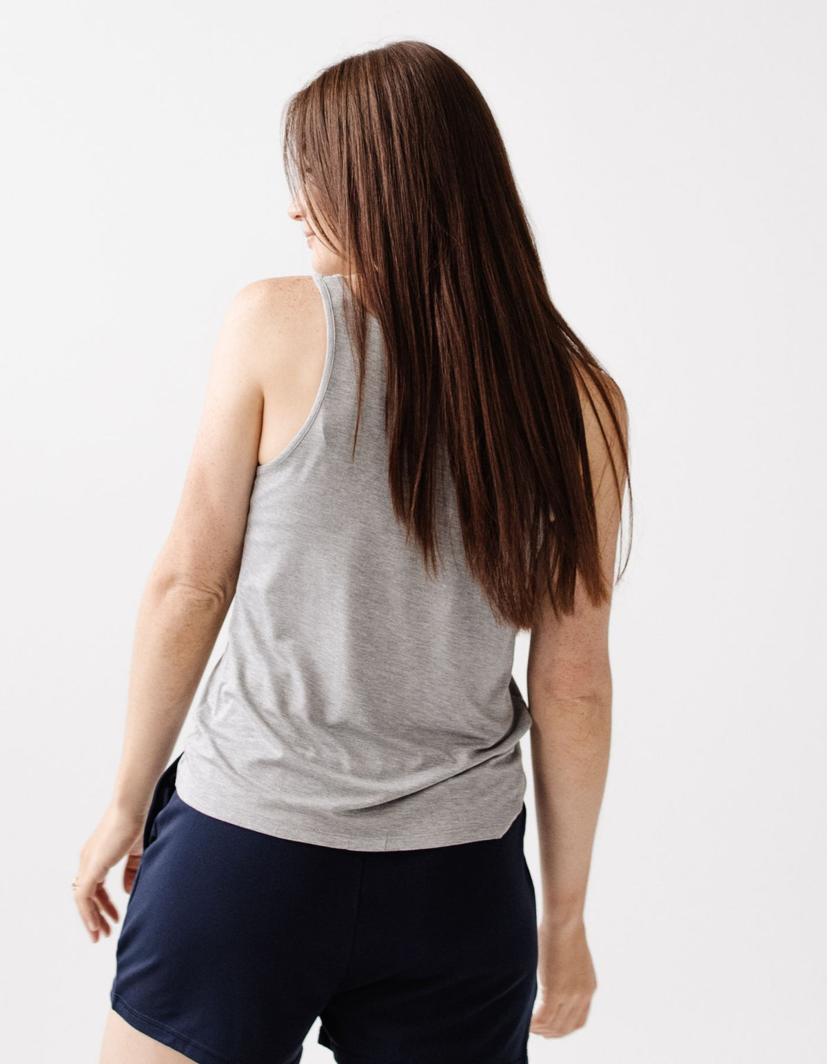 A woman with long brown hair wears the Cozy Earth Women's Bamboo Stretch-Knit Lounge Tank in gray, paired with navy shorts, standing against a plain white background and looking slightly over her shoulder.