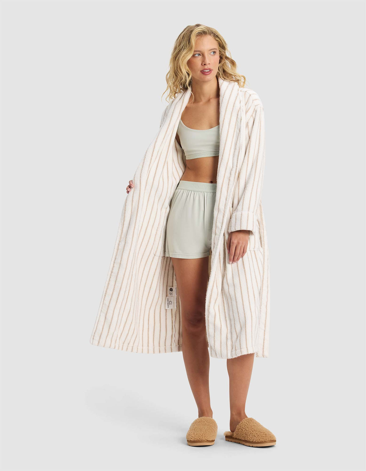 A woman stands against a plain light gray background, wearing the Cozy Earth Striped Luxe Bath Robe over a light green bralette and shorts set, paired with tan slippers.|Color:Sand Stripe