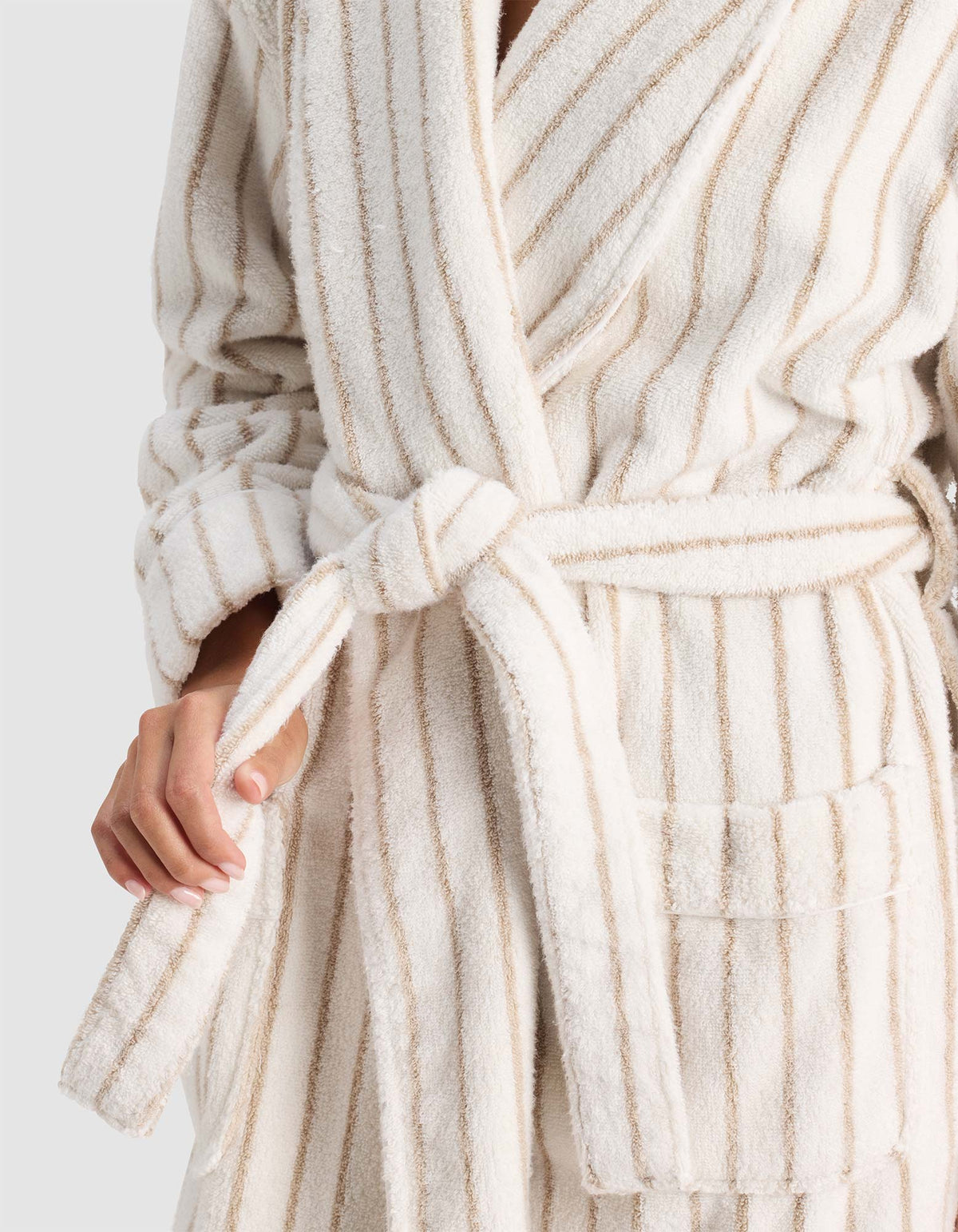 A person wears the Cozy Earth Striped Luxe Bath Robe, a soft cream-colored robe with beige vertical stripes, tied at the waist; only their upper body and one hand adjusting the belt are visible.