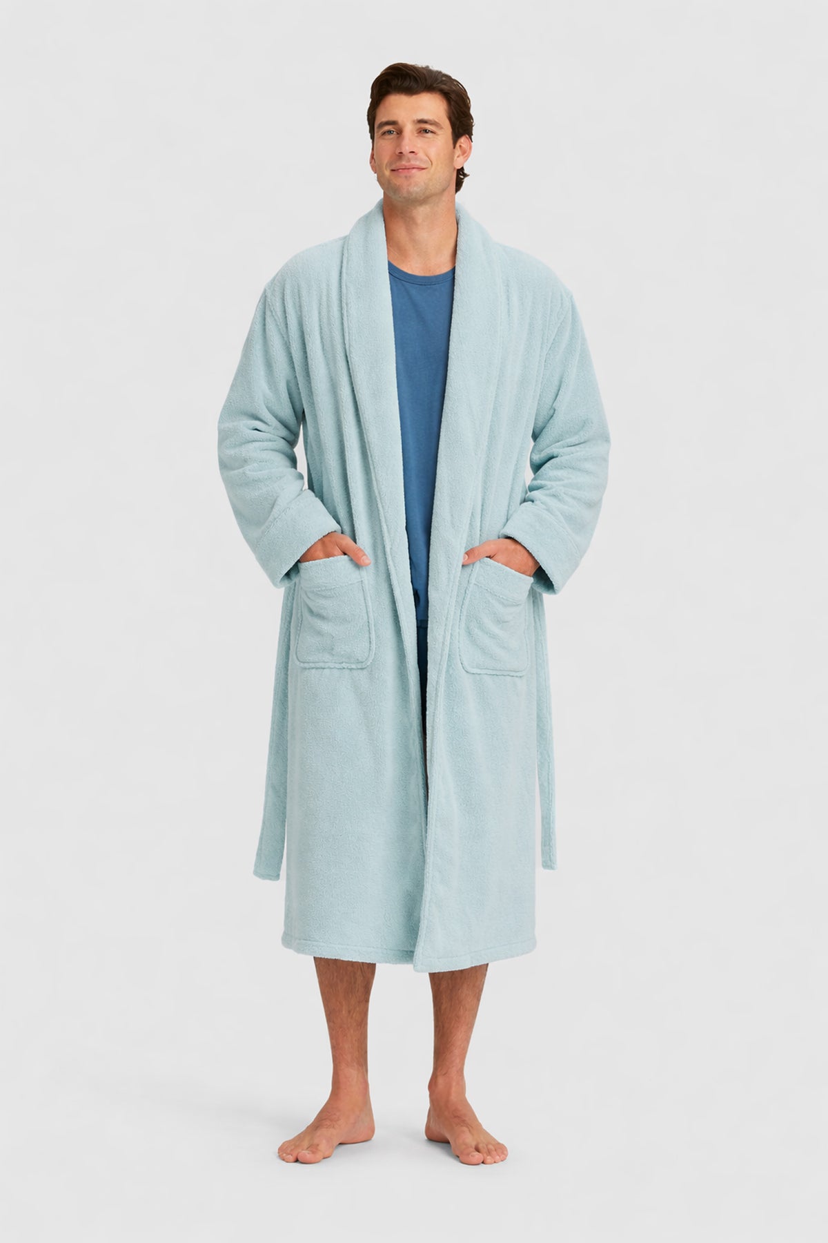 A man stands barefoot in a Luxe Bath Robe by Cozy Earth over a blue shirt, hands in the robe’s pockets, smiling gently against a plain light background.