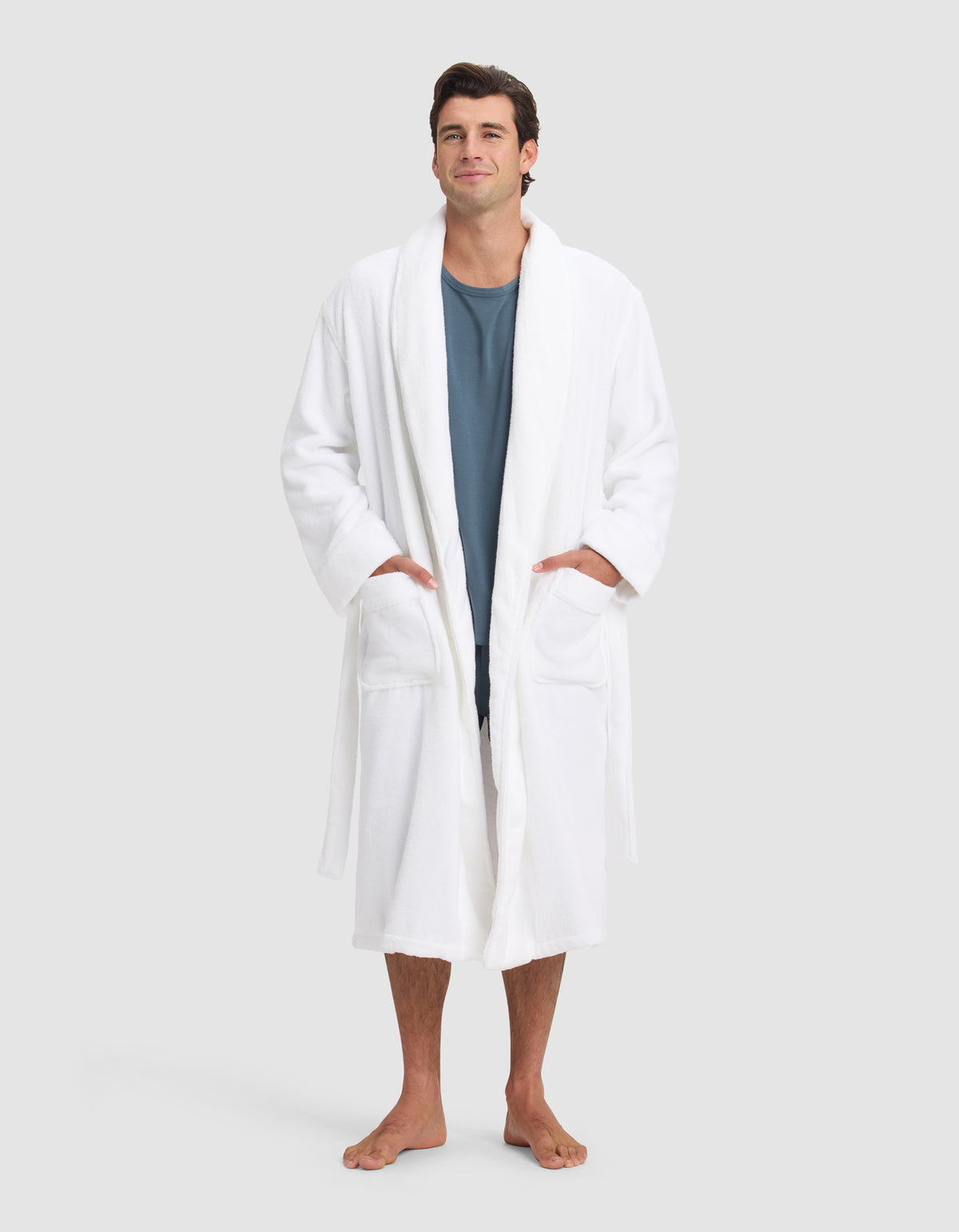 A man stands barefoot against a neutral background, wearing the Cozy Earth Luxe Bath Robe over a blue shirt. His hands are in the robe’s pockets as he looks forward with a slight smile.