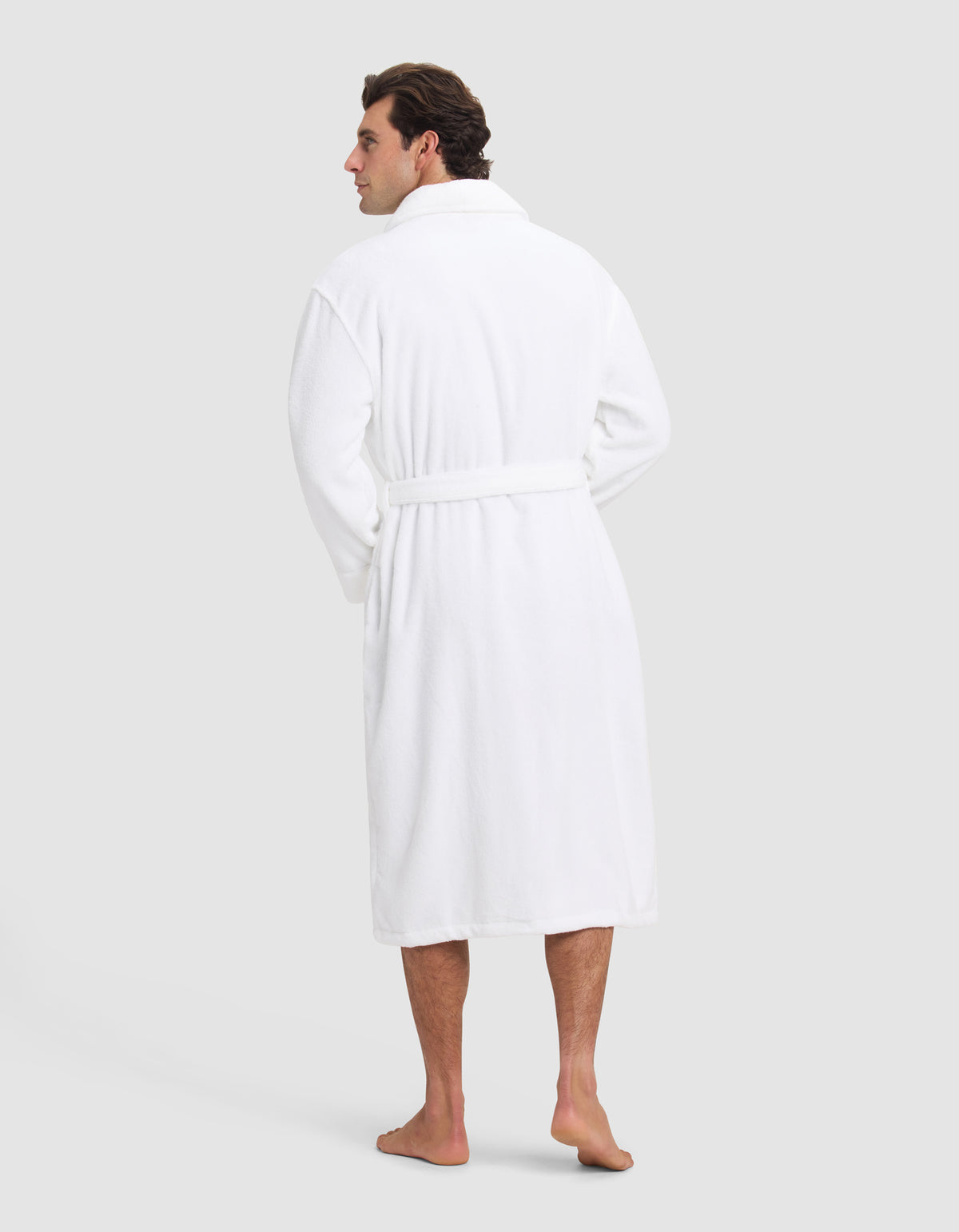 A man stands barefoot facing away, wearing the Luxe Bath Robe by Cozy Earth, featuring a belt, against a plain light gray background.