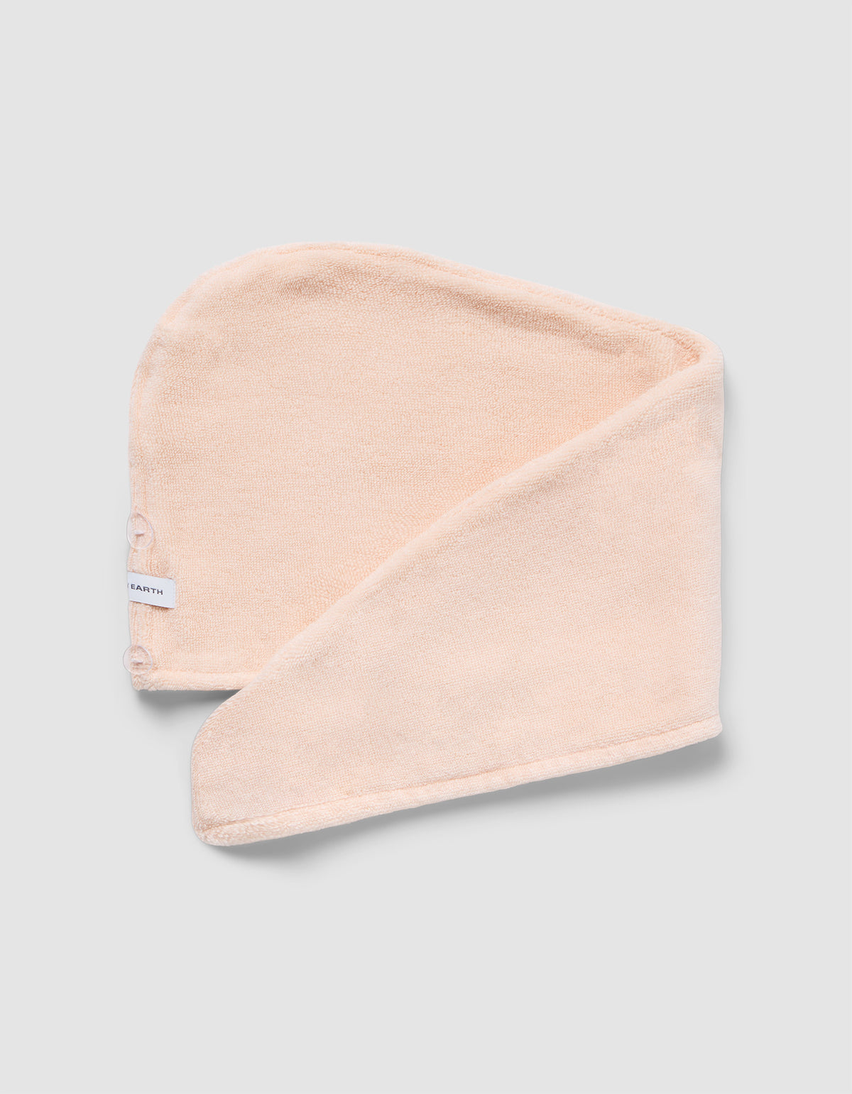 The Luxe Hair Towel by Cozy Earth is a soft, light beige microfiber towel with a white label and two snap buttons. Displayed partially folded on a light gray background, it highlights its rectangular shape and smooth texture.