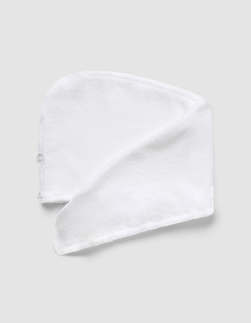 The Luxe Hair Towel by Cozy Earth, made of white microfiber with a button, is folded in a spiral and laid flat on a light gray background.